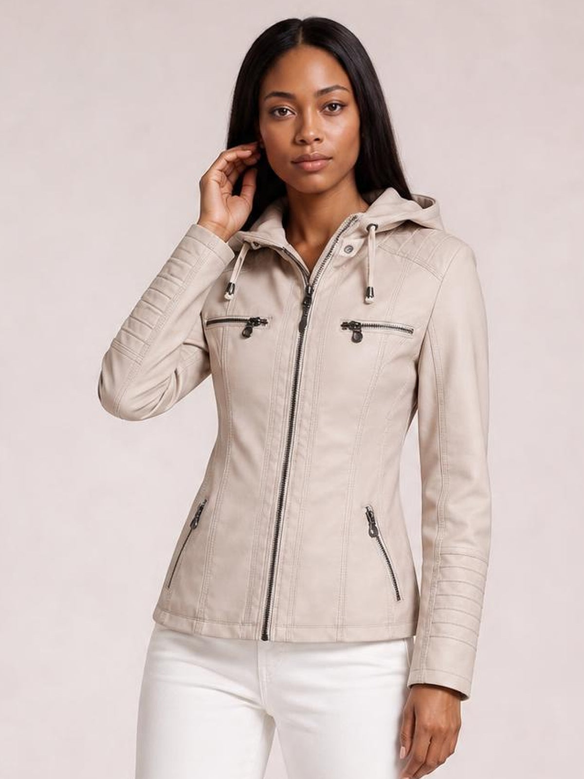 Model poses in a light beige faux leather moto jacket with a hood, zip closure, seam detailing, ribbed sleeves, and multiple zippered pockets.