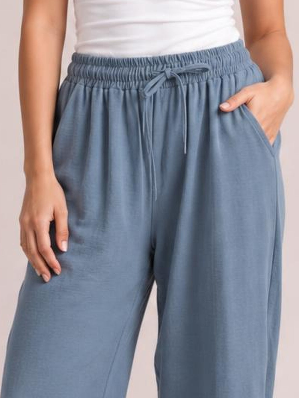 A model wears blue-gray drawstring cropped jogger pants with an elastic waistband, relaxed wide-leg silhouette, side pockets, and one hand tucked inside.