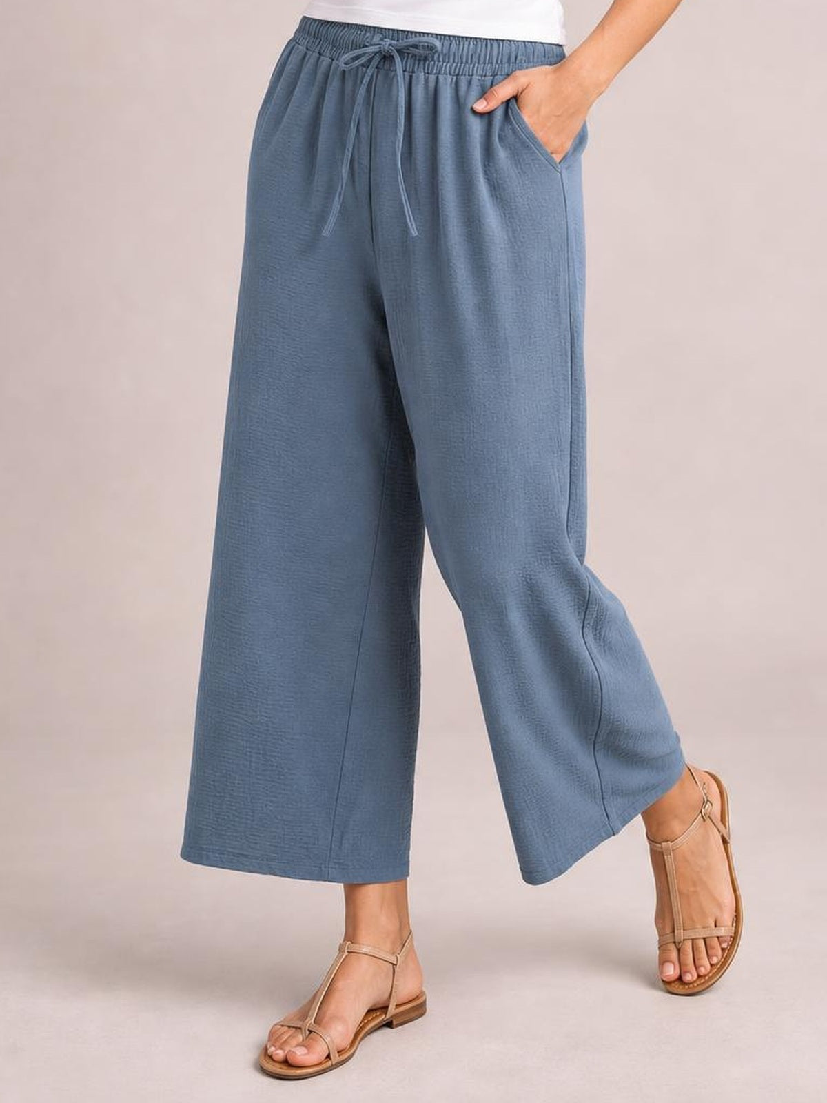 A model poses in blue-gray drawstring cropped jogger pants with an elastic waist, wide-leg silhouette, side pockets, and one hand tucked into a pocket.