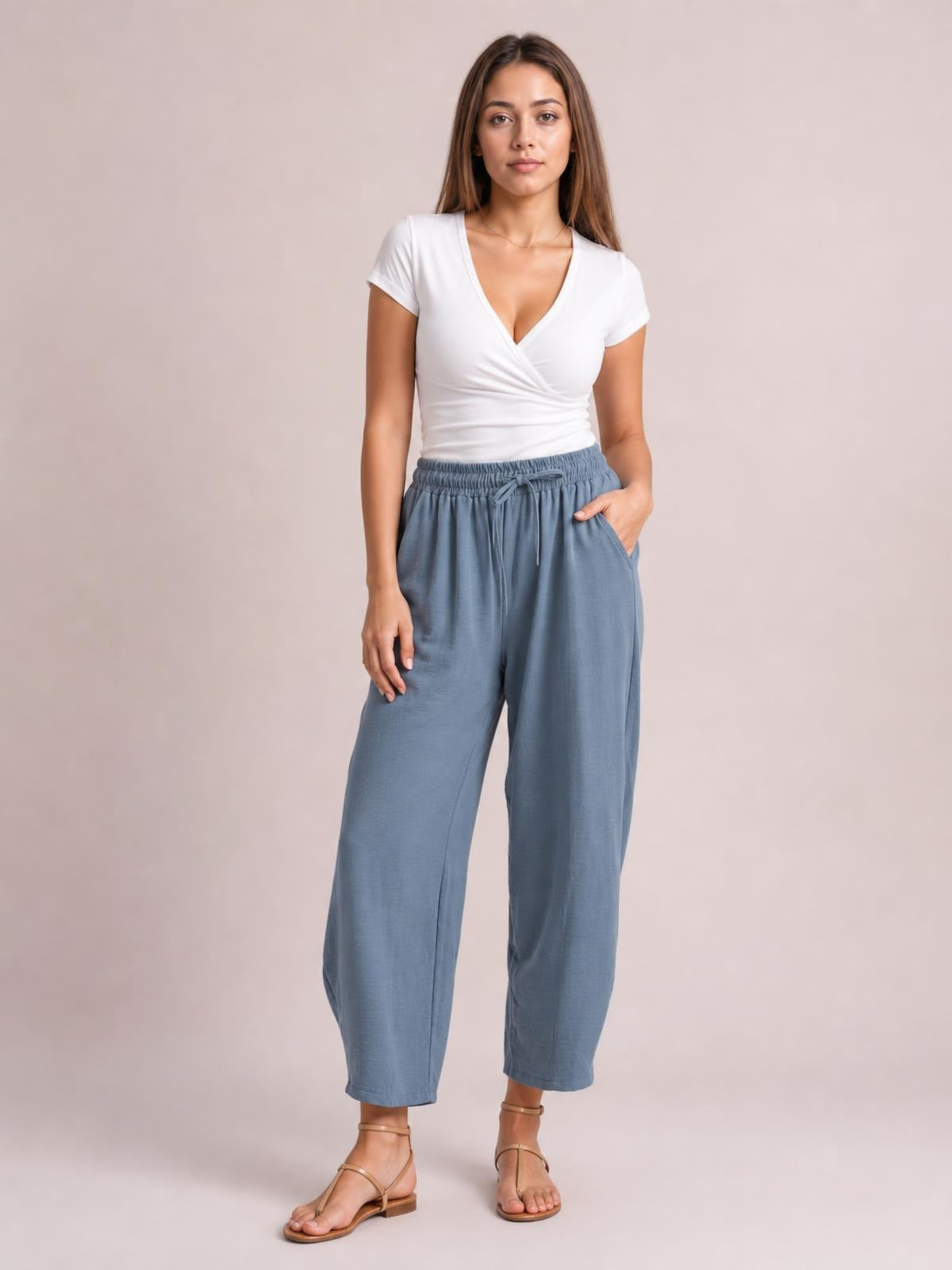 A model stands in blue-gray cropped jogger pants with a gathered drawstring waist, relaxed wide-leg silhouette, and side pockets, resting one hand in a pocket.
