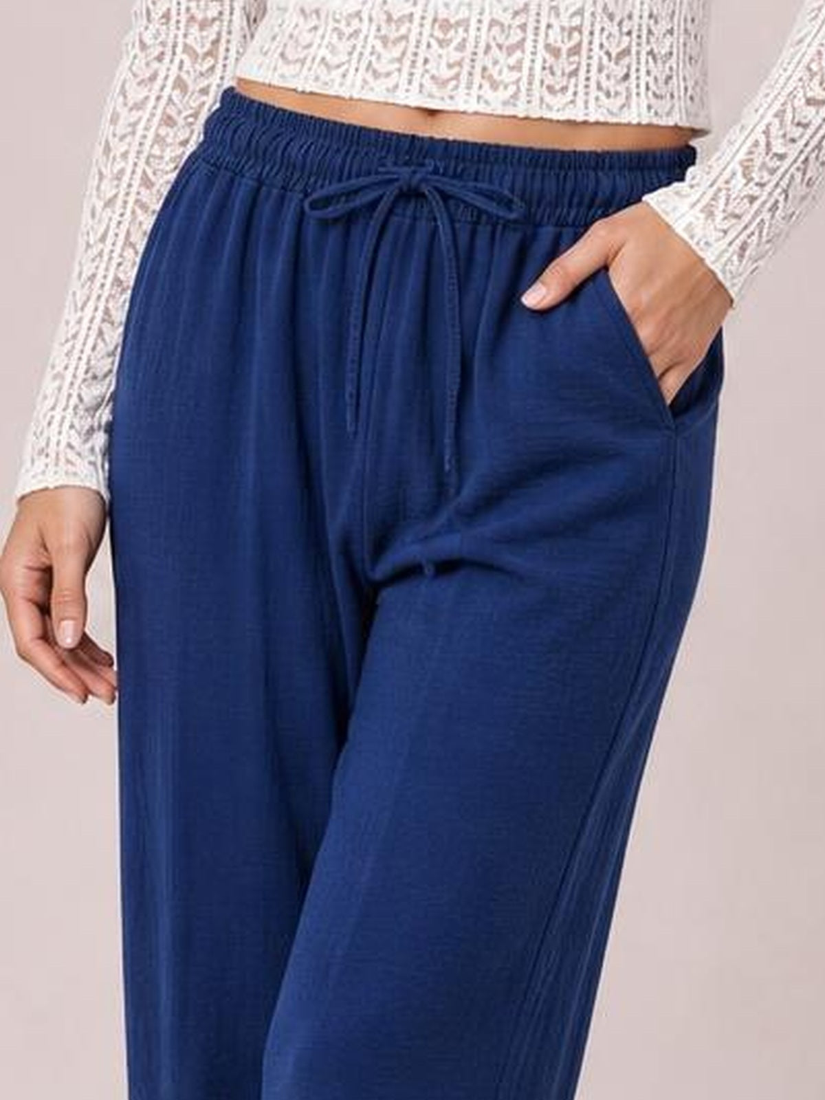 A model wears navy blue cropped jogger pants with a gathered drawstring waistband, relaxed silhouette, and side pockets, posing with one hand tucked inside.