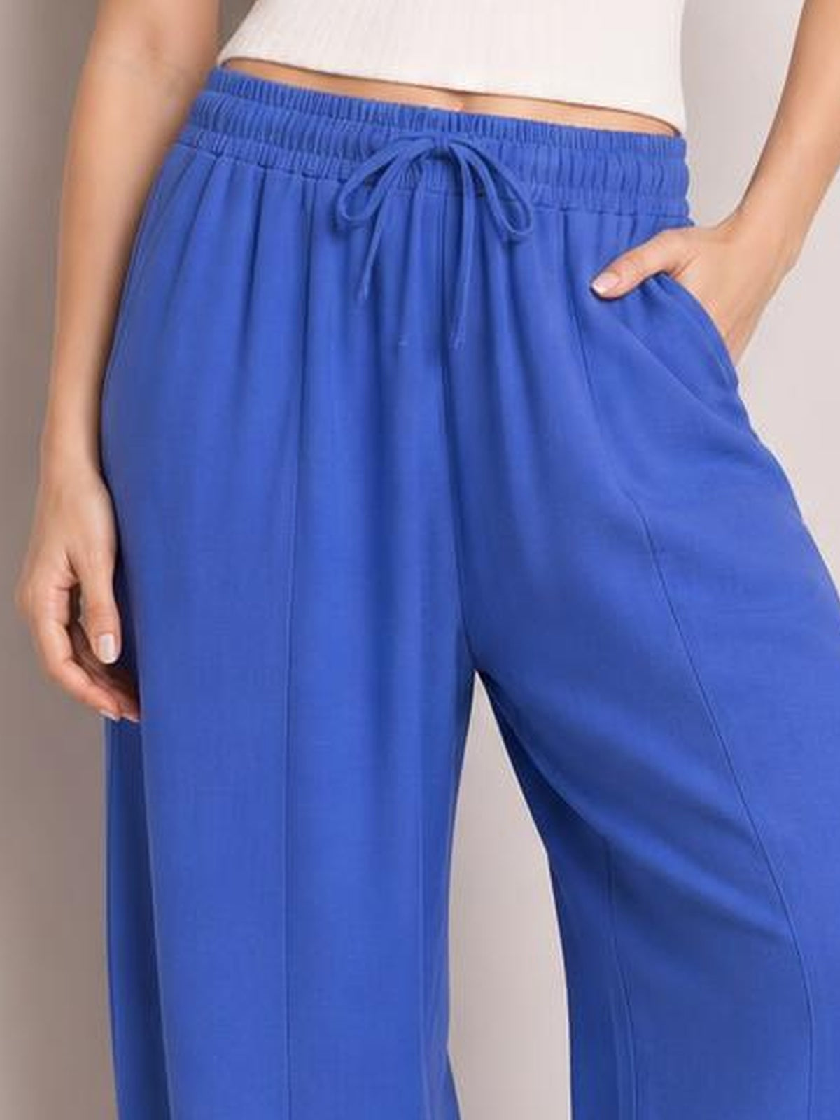 A model wears bright blue drawstring cropped jogger pants with an elastic waistband, relaxed wide-leg silhouette, and side pockets, posing with one hand tucked in.