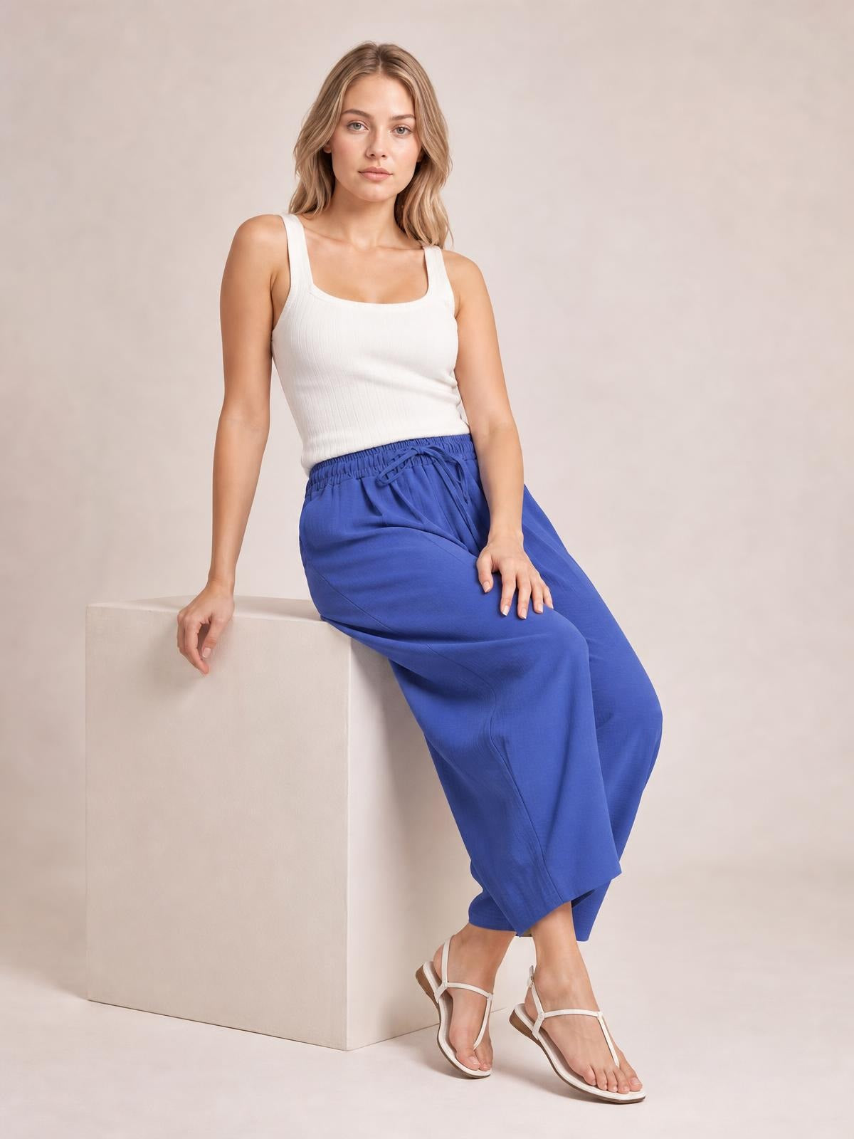 A seated model wears bright blue drawstring cropped jogger pants with an elastic waist, relaxed tapered silhouette, and softly structured construction.