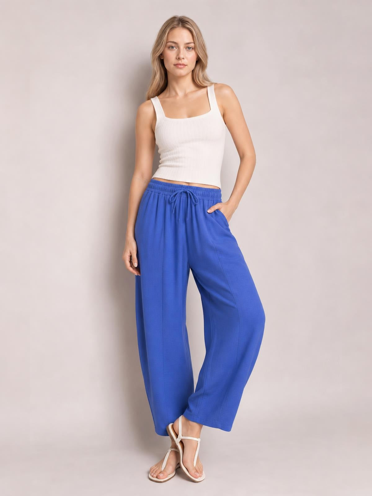 A model poses in bright blue cropped jogger pants with a drawstring elastic waist, relaxed wide-leg silhouette, and side pockets, one hand tucked in a pocket.