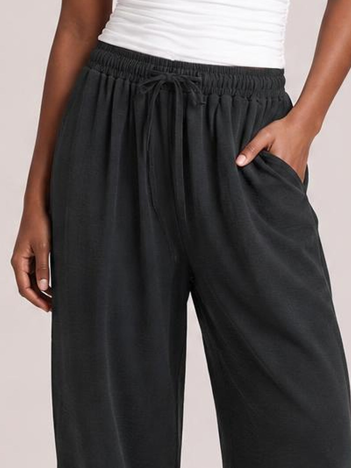 Model poses with one hand in a pocket, wearing black cropped jogger pants with a drawstring elastic waist and relaxed wide-leg silhouette.
