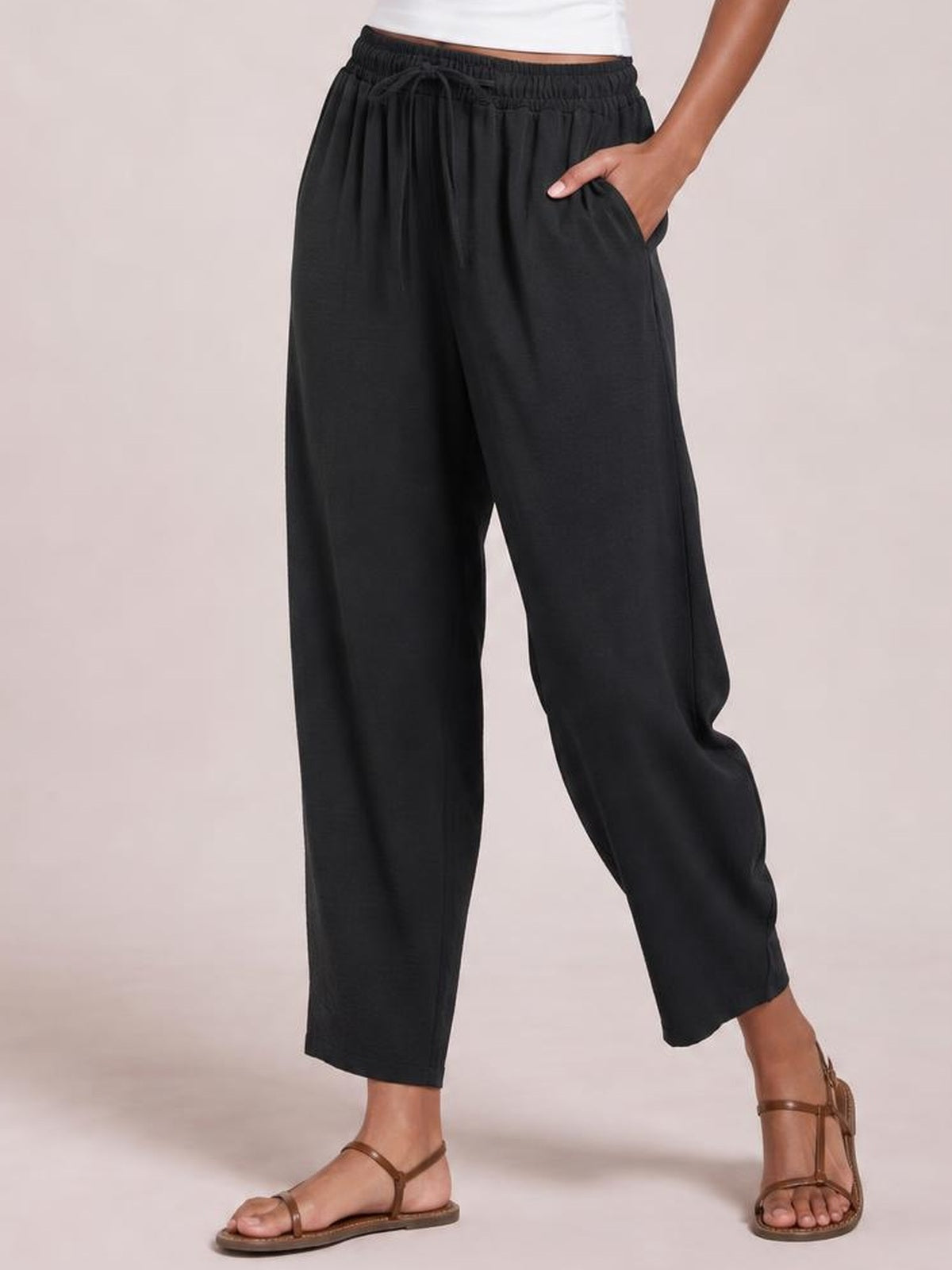 Model wears black drawstring cropped jogger pants with an elastic waist, wide relaxed legs, side pockets, and ankle-length hems, standing with one hand in a pocket.