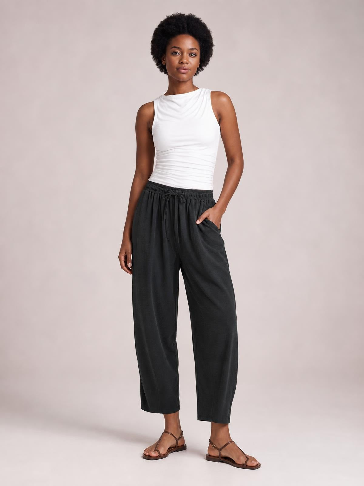 A model stands in black cropped jogger pants with a drawstring waistband, roomy silhouette, visible side pockets, and one hand tucked into a pocket.