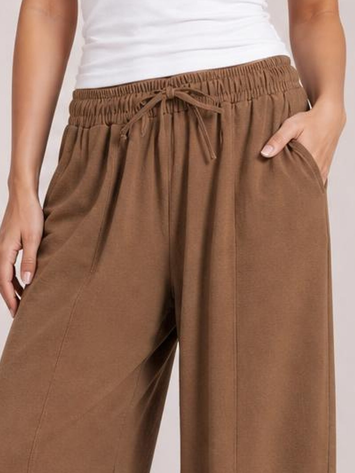 A model wears brown drawstring cropped jogger pants with an elastic waistband, relaxed wide-leg silhouette, visible front pockets, and hands tucked inside.