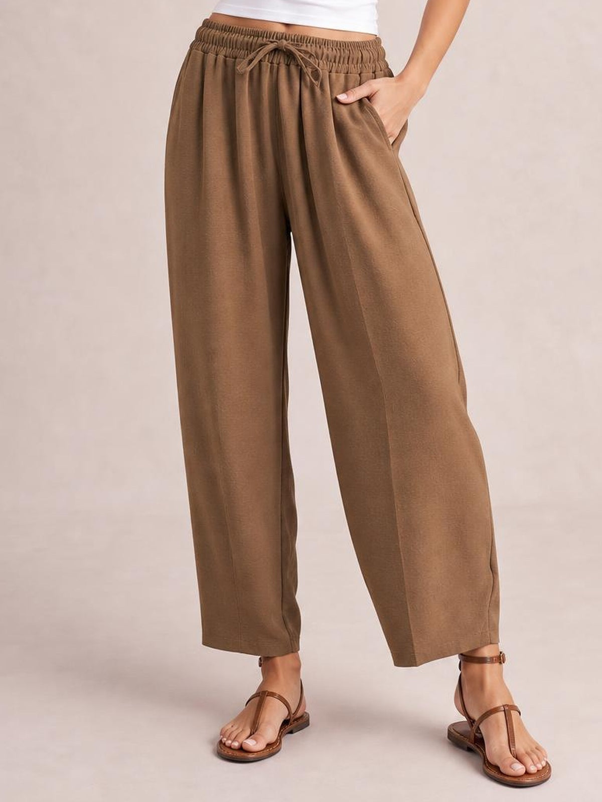 A model wears brown cropped jogger pants with a gathered drawstring waist, relaxed wide-leg silhouette, side pockets, and one hand tucked into a pocket.