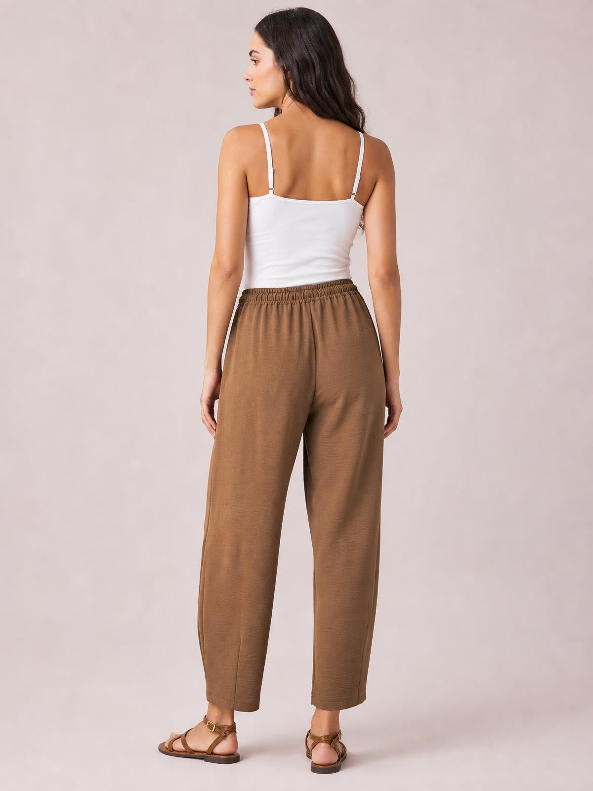 A model stands turned away in brown drawstring cropped jogger pants with an elastic waist and relaxed, straight-leg silhouette ending above the ankles.