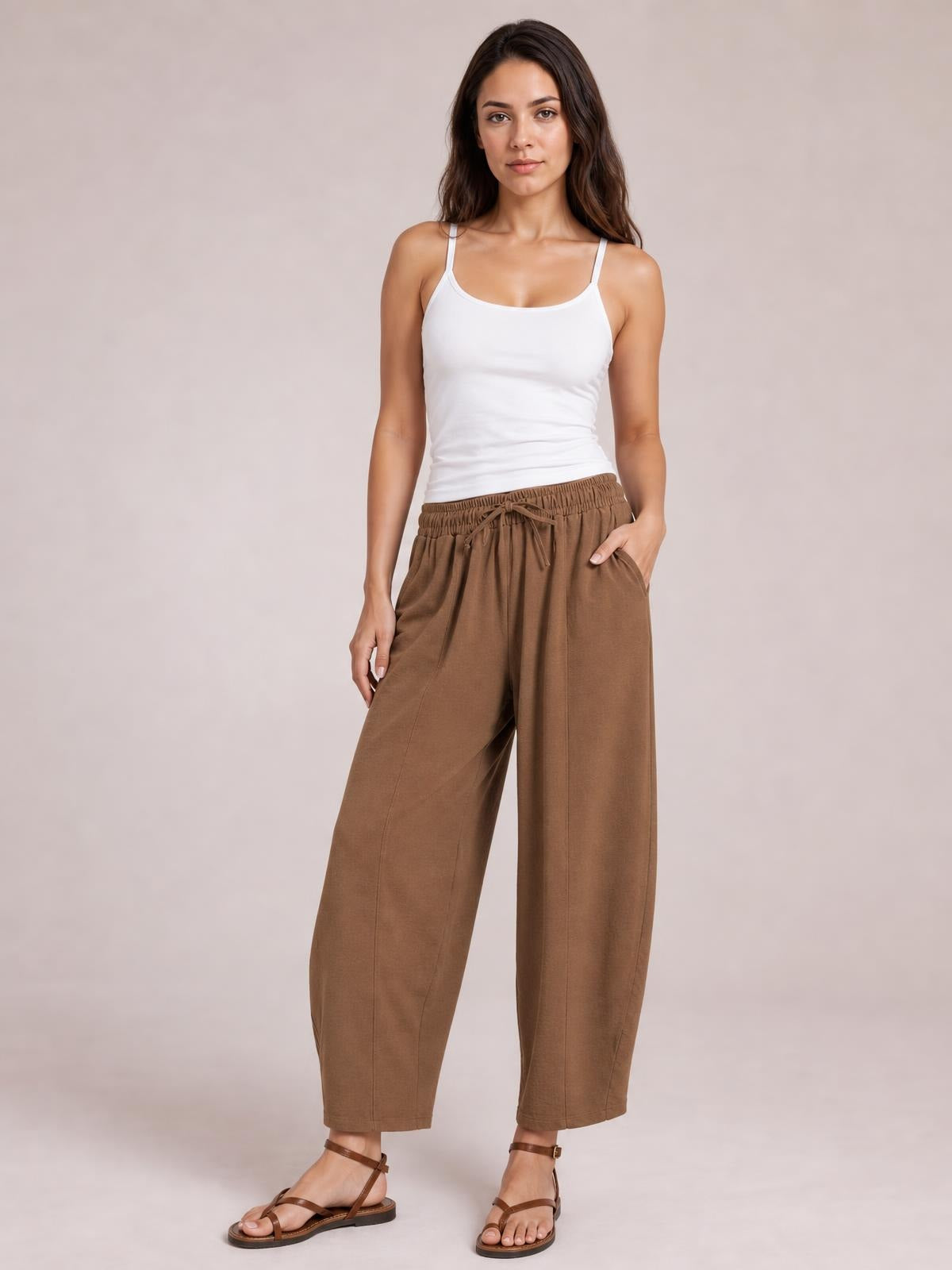 Model poses with one hand in a pocket, wearing brown drawstring cropped jogger pants with an elastic waist, relaxed wide-leg silhouette, and ankle-length hems.