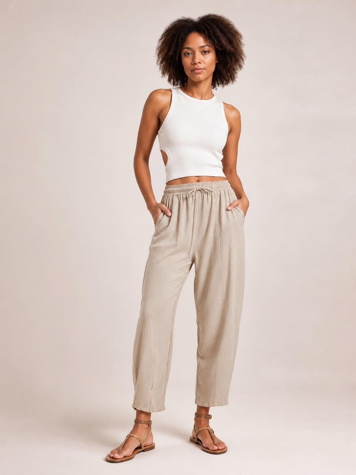 A model wears light beige drawstring cropped jogger pants with an elastic waistband, visible pockets, and a relaxed tapered silhouette while standing with hands tucked inside.