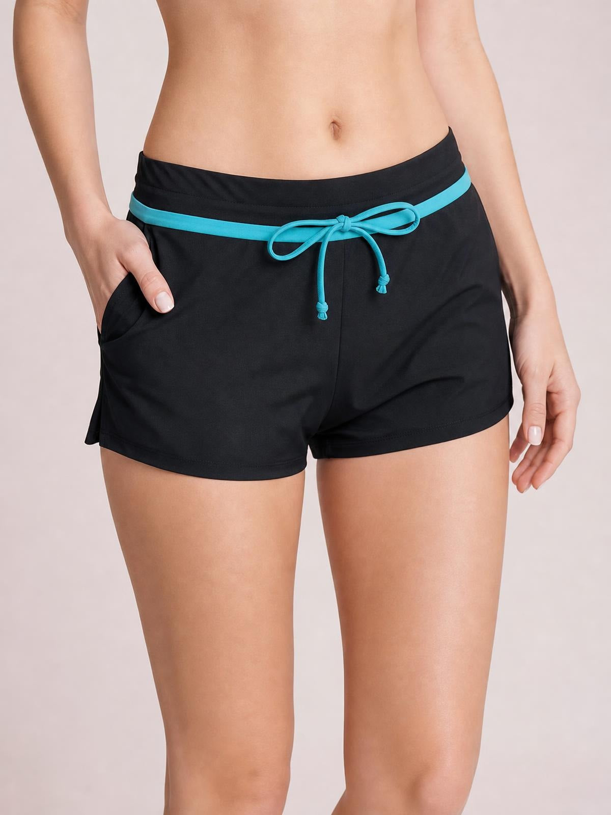 Model wearing black drawstring swim shorts with a relaxed short-leg silhouette, turquoise waistband trim and front tie, with one hand in a side pocket.