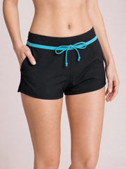 Model wearing black drawstring swim shorts with a relaxed short-leg silhouette, turquoise waistband trim and front tie, with one hand in a side pocket.