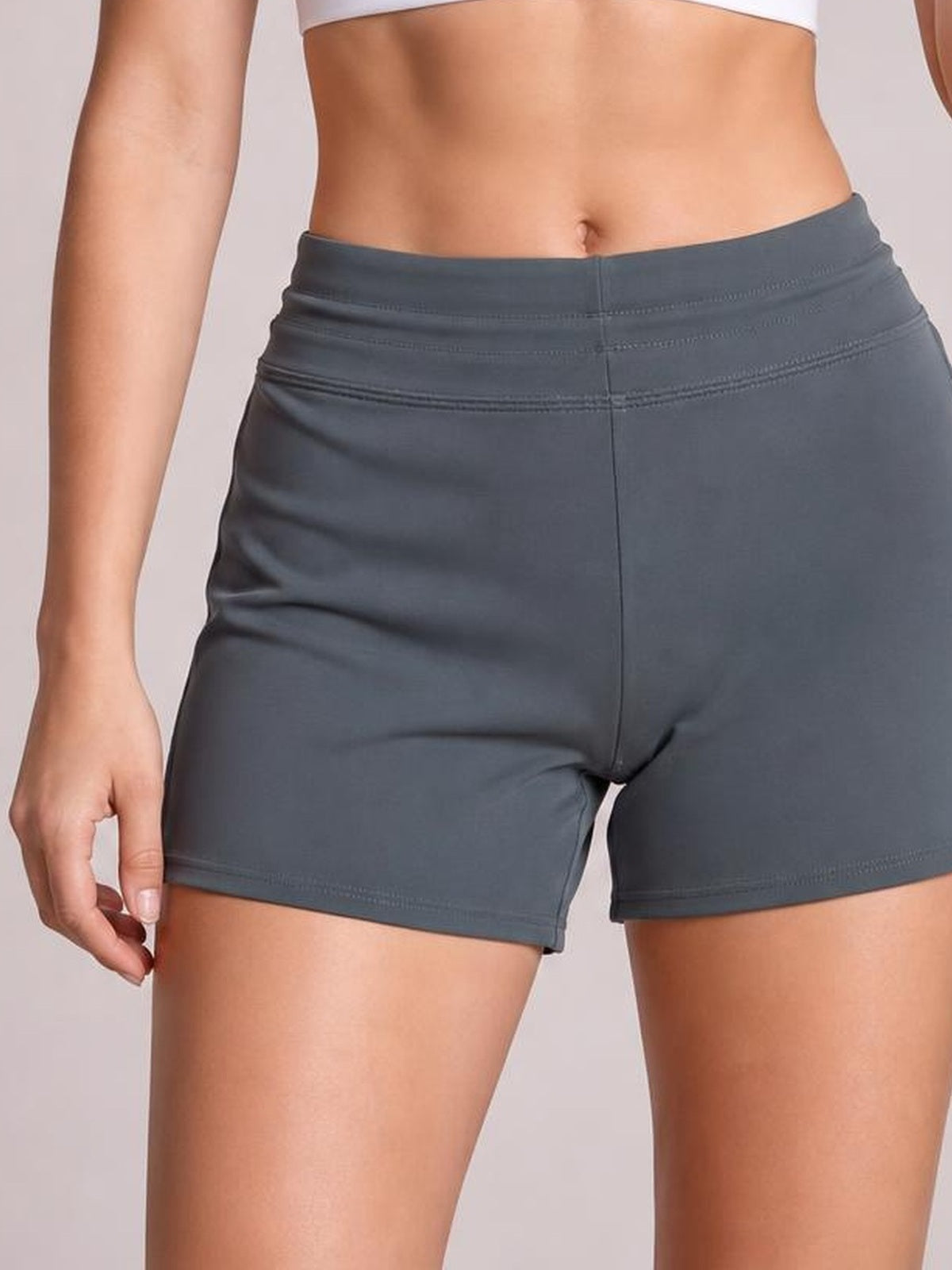 A cropped model wears charcoal gray drawstring swim shorts with a stretchy fitted silhouette, seamed waistband, and straight hems while lightly touching one leg.