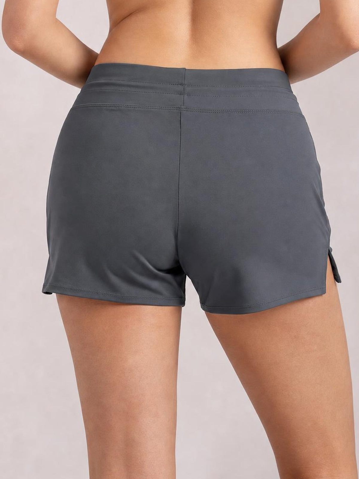 A rear view of a model wearing dark gray drawstring swim shorts with a stretchy, fitted silhouette, elastic waistband, side slit, and stitched hems.