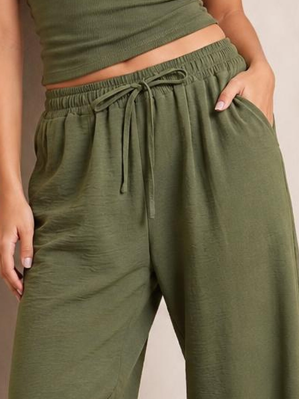 A model wears olive green jogger pants with a relaxed wide-leg silhouette, elastic drawstring waistband, side pockets, and one hand tucked into a pocket.