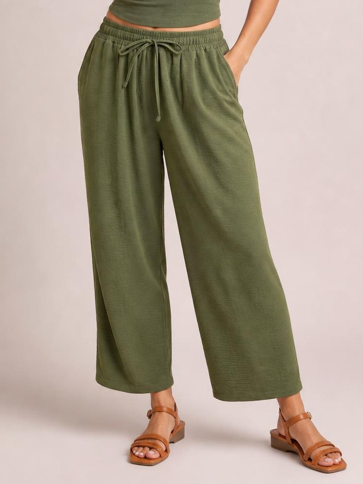 A model wears olive green cropped jogger pants with an elastic drawstring waist, wide-leg silhouette, and side pockets, posing with one hand tucked in.