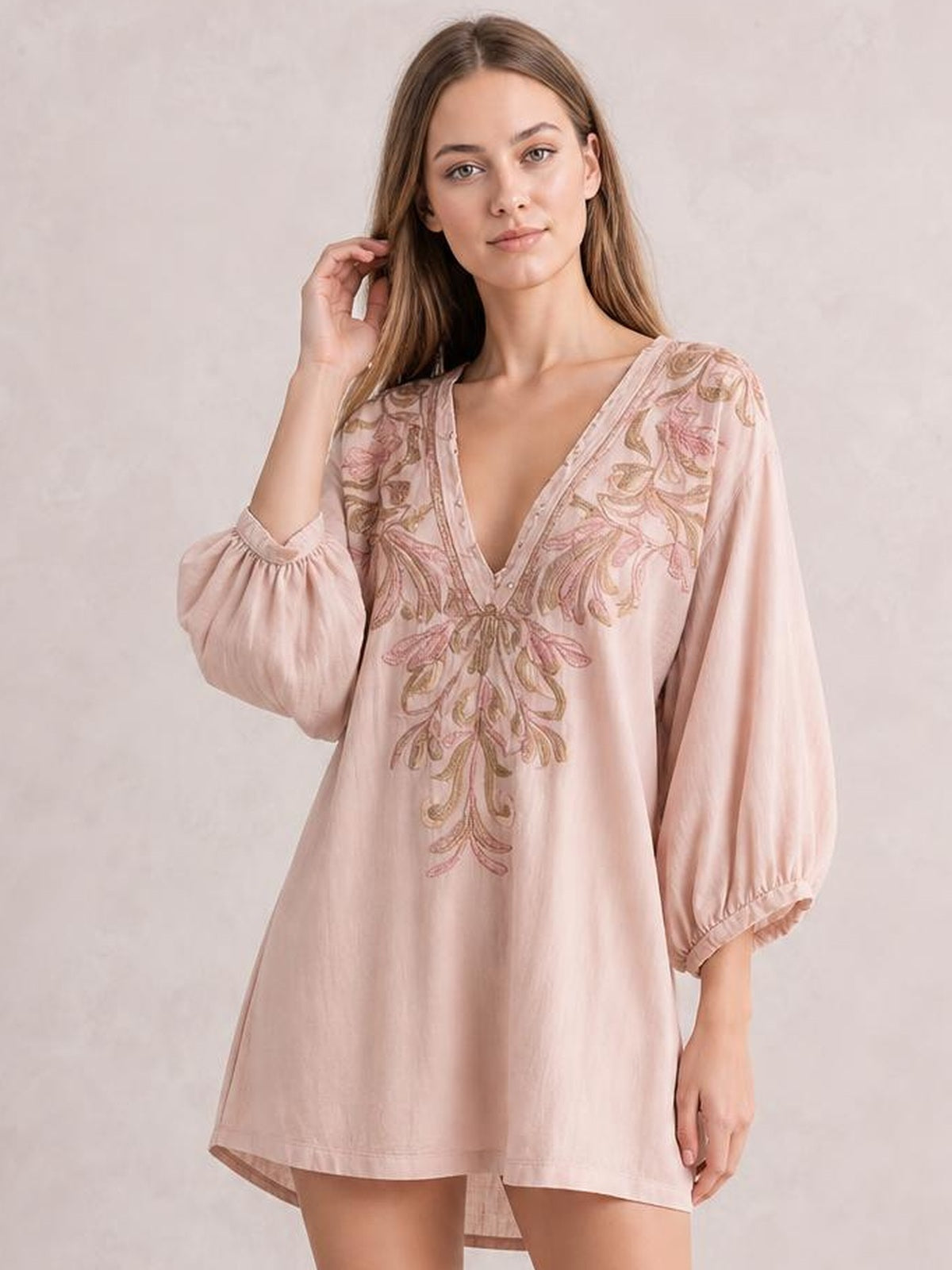 A model poses in a pale pink embroidered V-neck mini dress with a loose A-line silhouette, balloon sleeves, and elastic cuffs, touching her hair.