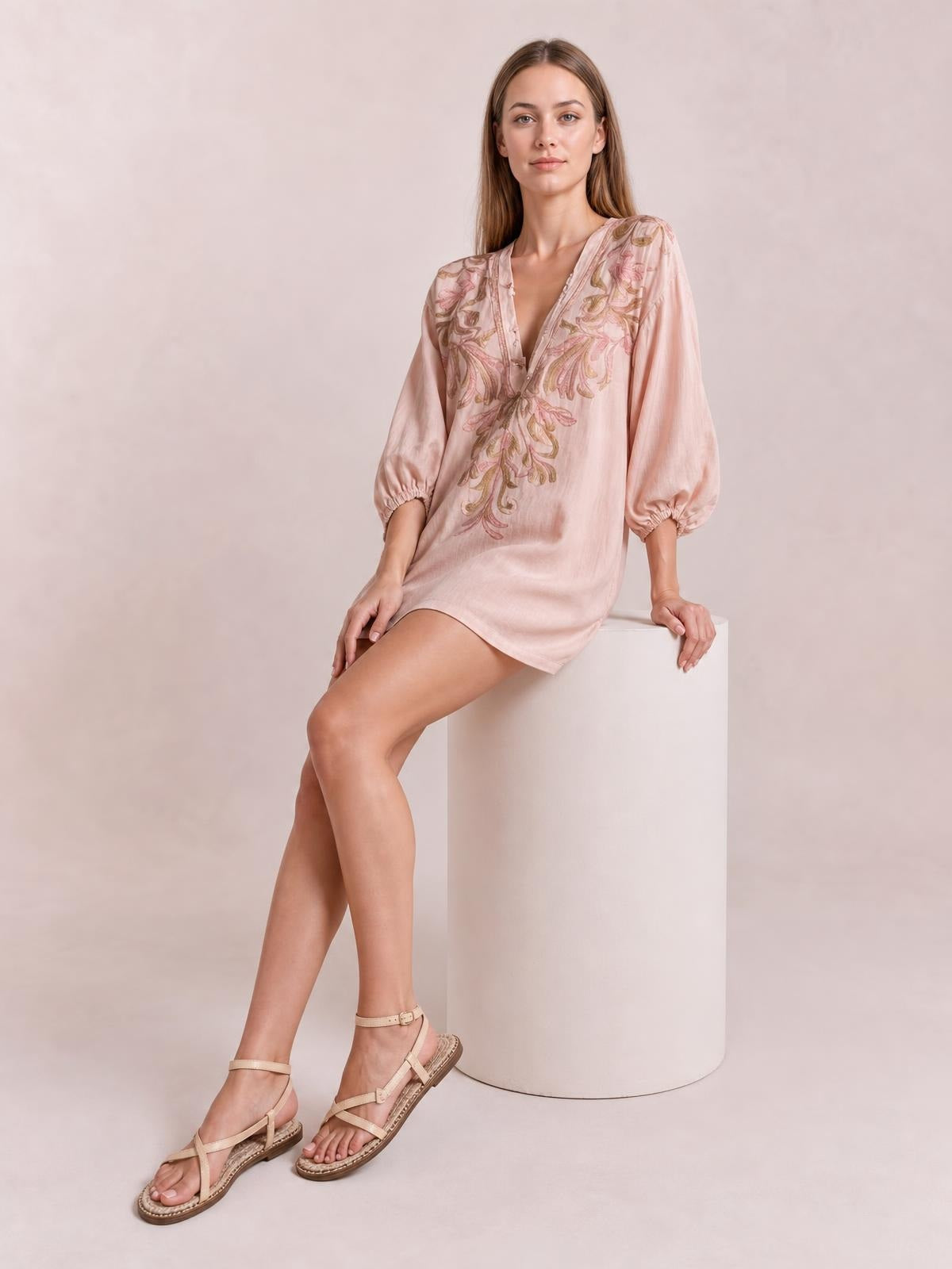 A model sits wearing a pale pink embroidered V-neck mini dress with a relaxed silhouette, floral detailing, and voluminous three-quarter sleeves with elastic cuffs.