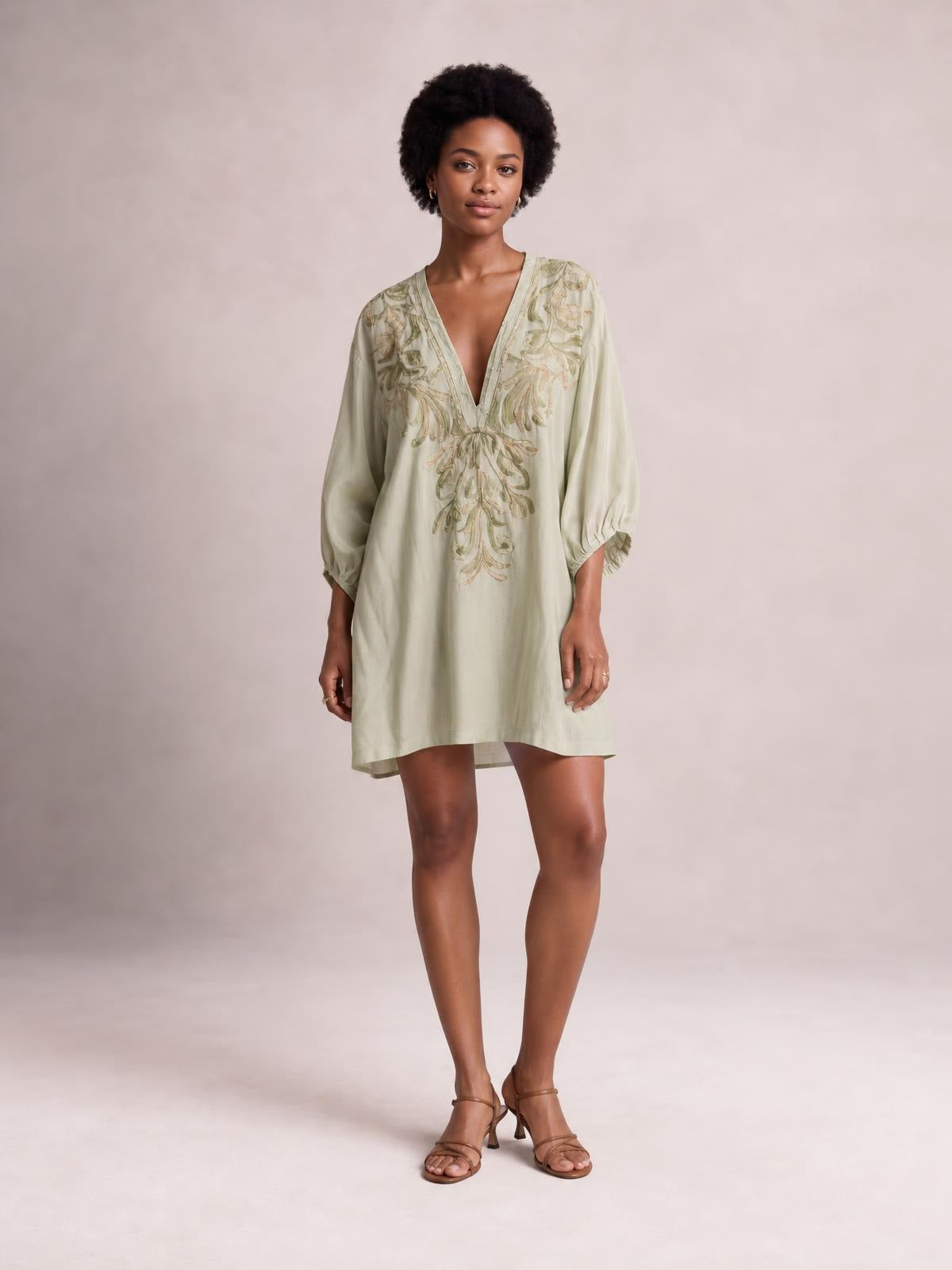 A model stands in a pale sage embroidered V-neck mini dress with a relaxed silhouette, flowing three-quarter sleeves, and softly gathered cuffs.