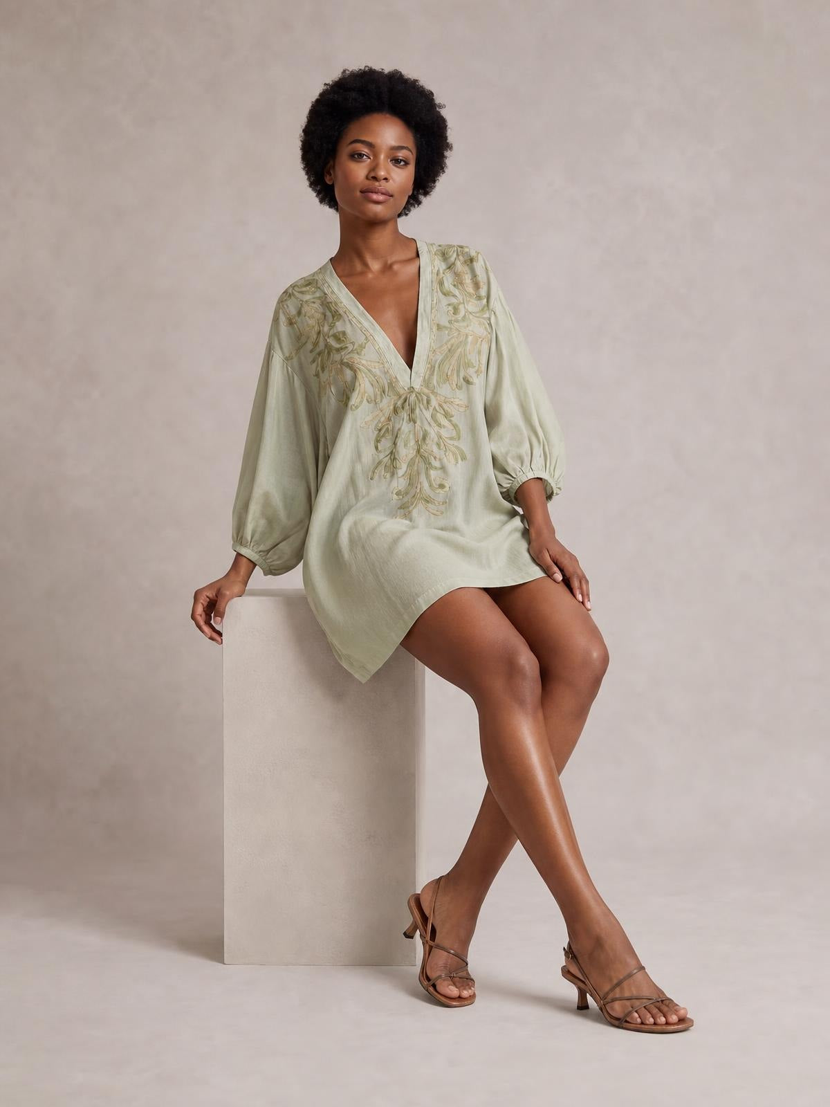 A model sits cross-legged in a pale sage embroidered V-neck mini dress with a relaxed silhouette, voluminous long sleeves, and gathered cuffs.