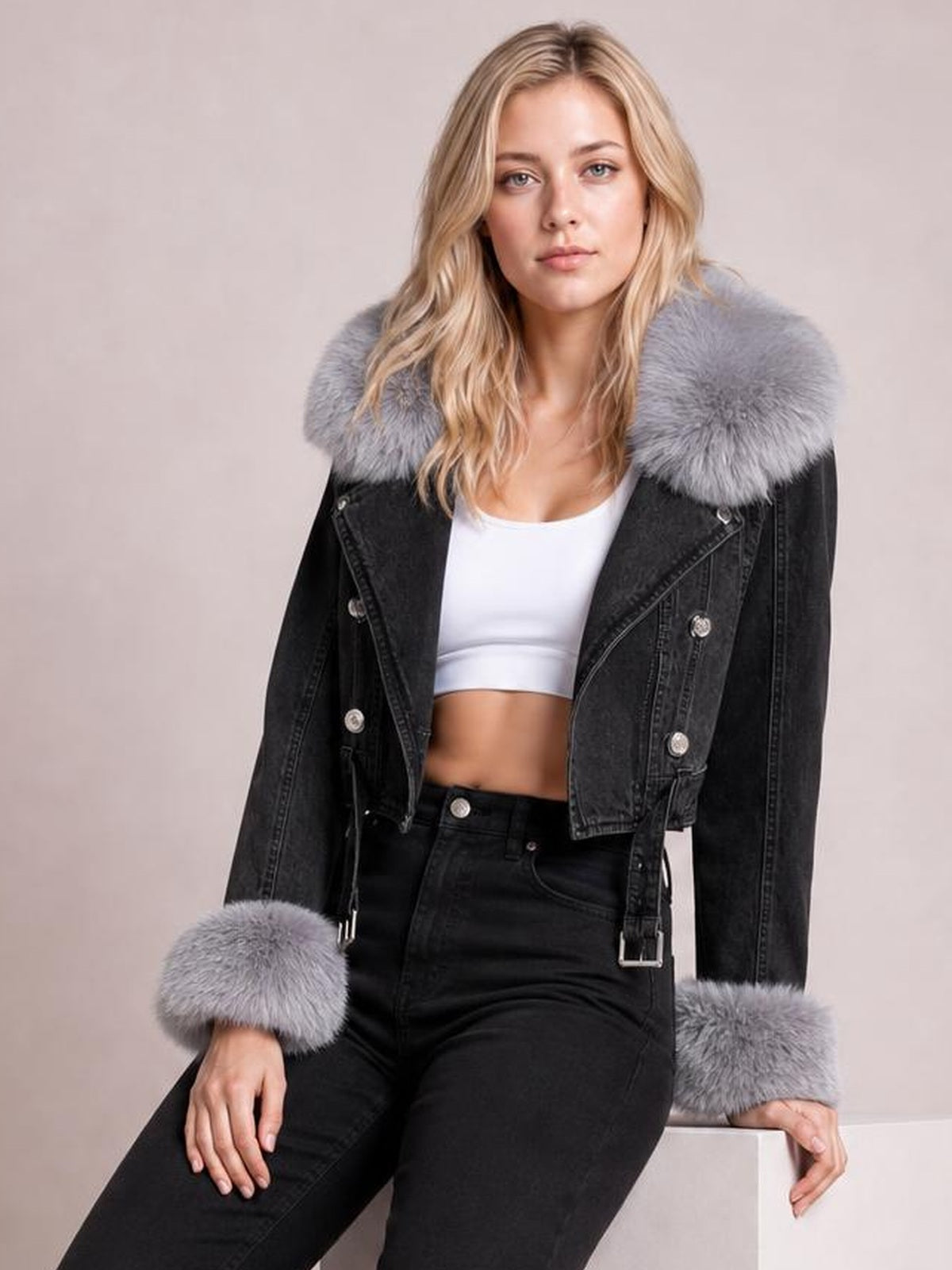 A seated model wears a black denim jacket with a cropped silhouette, double-breasted buttons, gray faux fur collar and cuffs, and belted waist details.