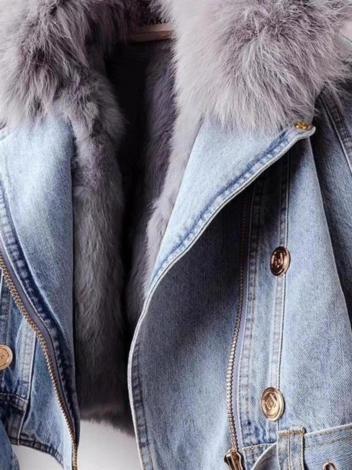 Close-up of a light blue denim jacket with a plush gray faux fur collar, front zipper, gold-tone buttons, and structured seamed construction.