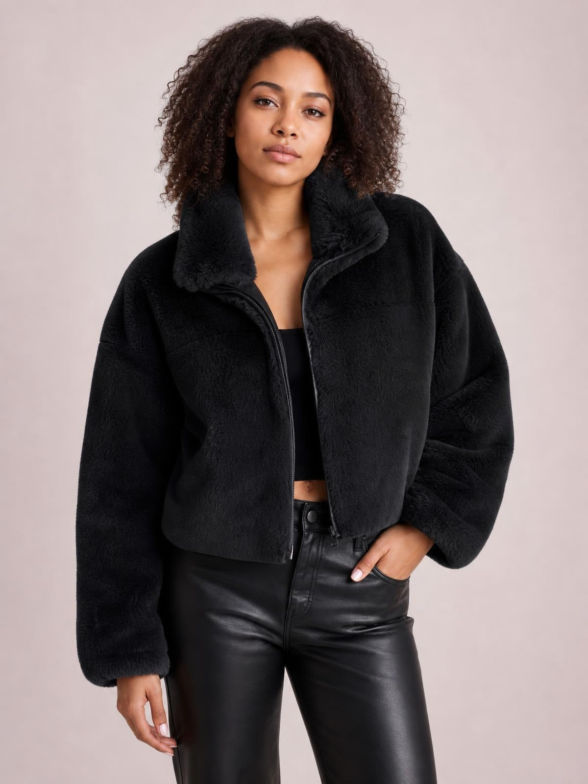 A model poses in a black faux fur cropped zip-up jacket with an oversized silhouette, plush high collar, long sleeves, and visible front zipper.