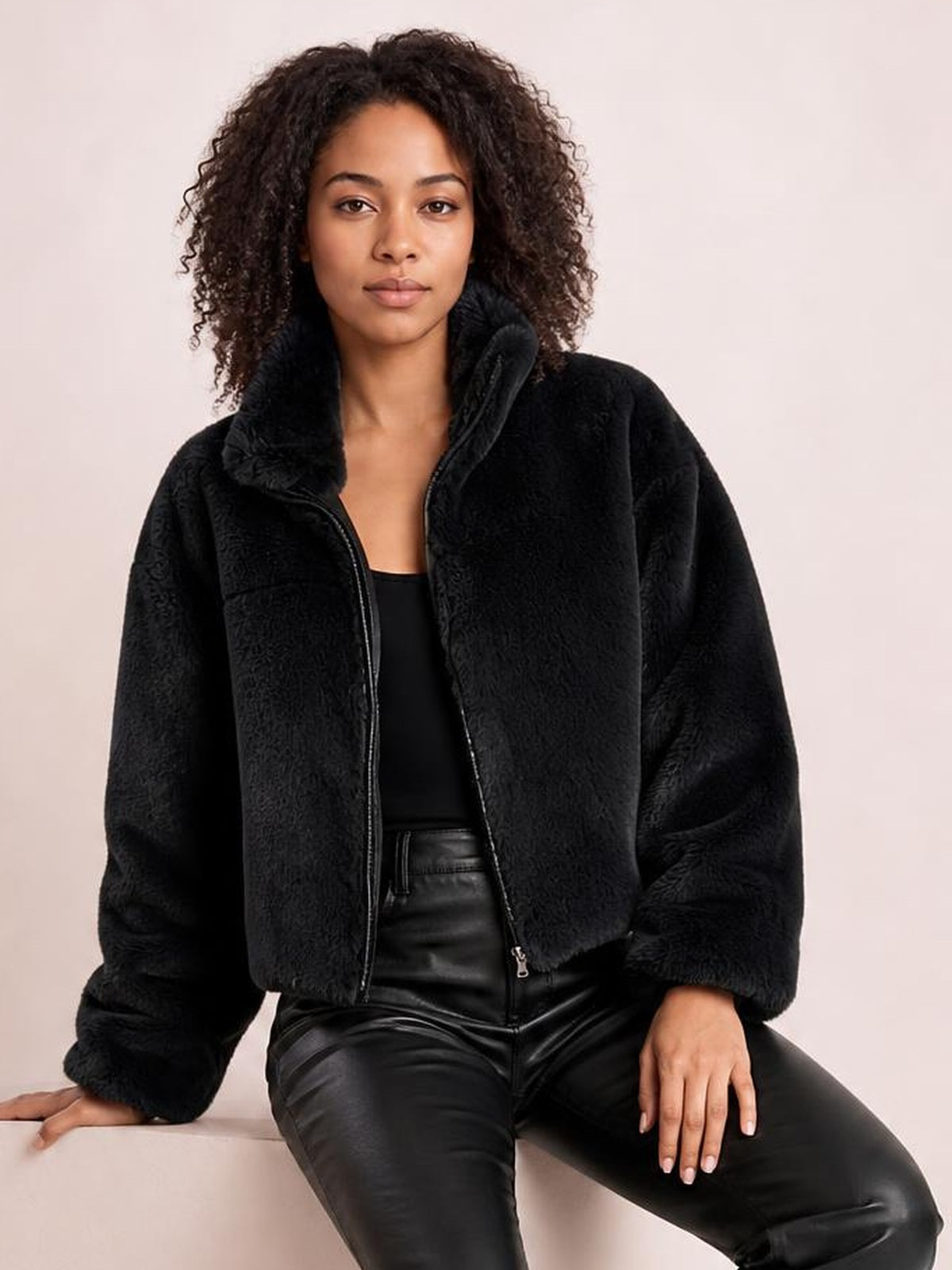 A seated model wears a black faux fur cropped zip-up jacket with an oversized silhouette, plush texture, raised collar, and visible front zipper.