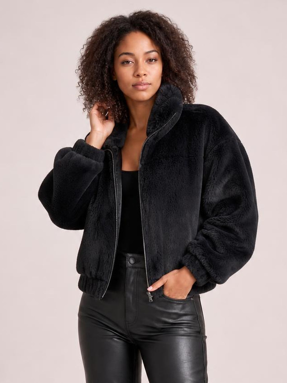 Model poses with one hand in her pocket wearing a black oversized faux fur cropped zip-up jacket with a high collar, thick texture, and elastic cuffs.