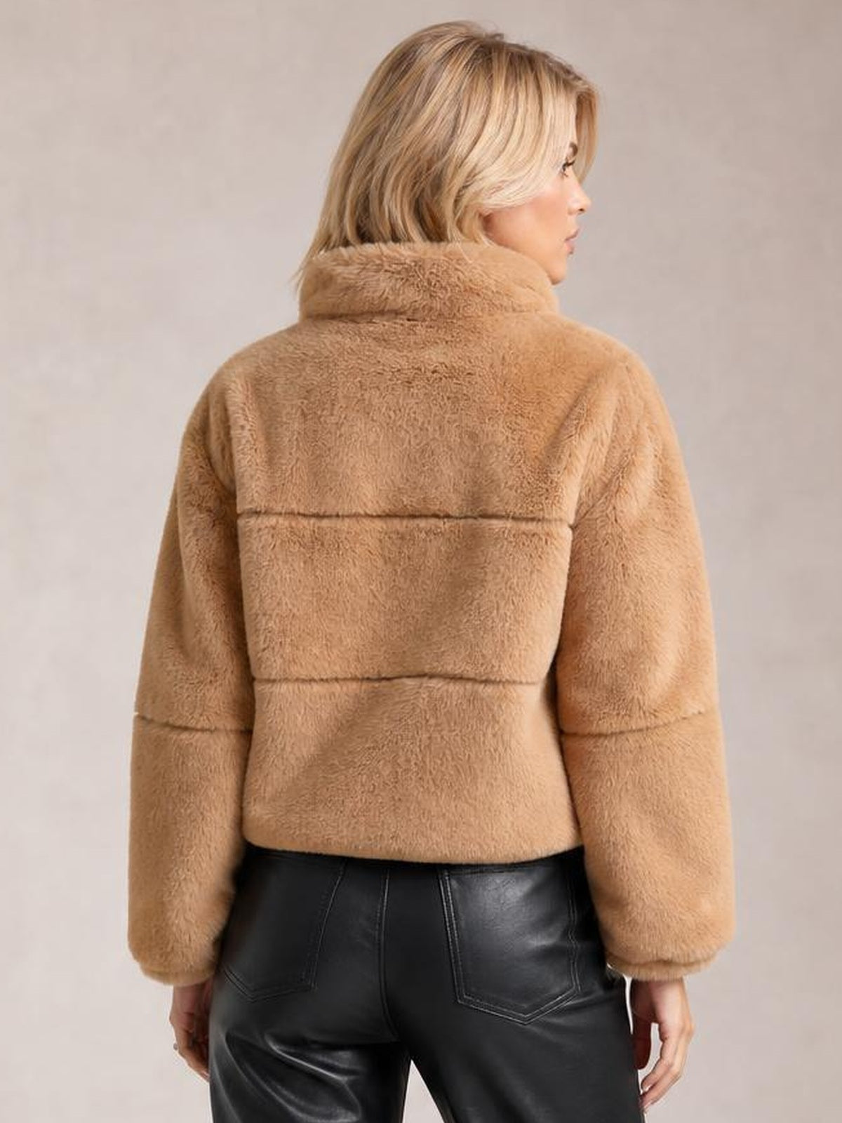 Model looks over her shoulder in a camel faux fur cropped zip-up jacket with an oversized silhouette, plush high collar, long sleeves, and horizontal seam detailing.