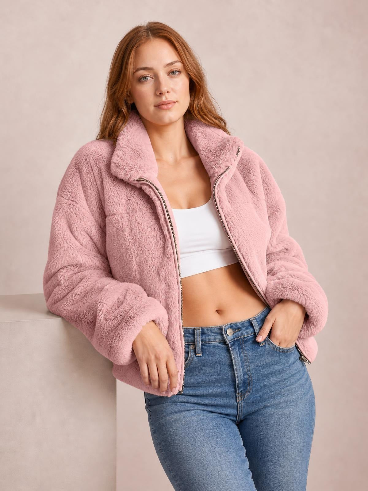 Model wears a pink faux fur cropped zip-up jacket with an oversized silhouette, plush texture, wide collar, and long sleeves, posing with one hand in her jeans pocket.