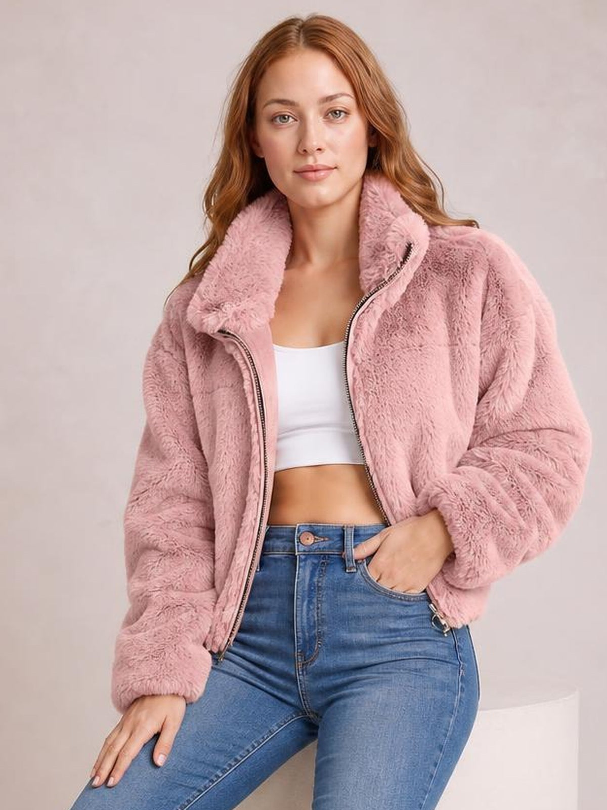 A model sits in a pink oversized faux fur cropped zip-up jacket with a plush high collar, open zipper, and voluminous sleeves, one hand in her jeans pocket.