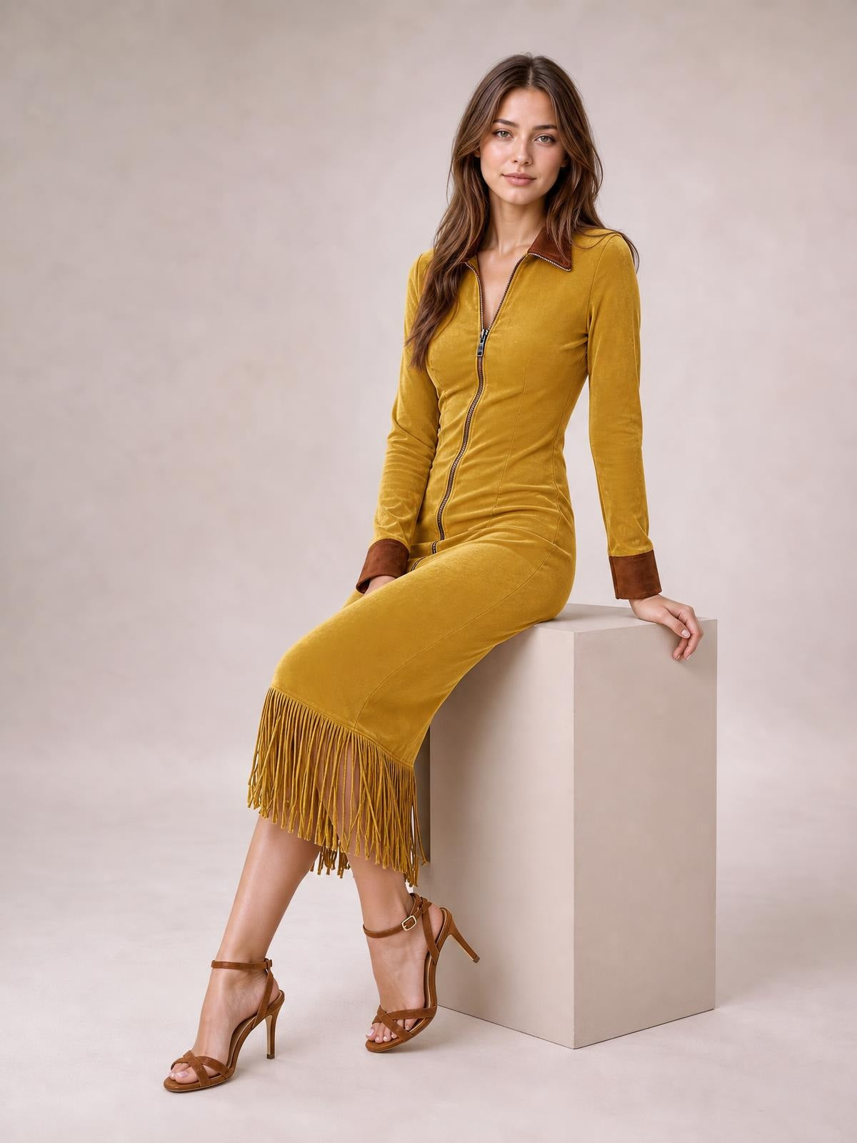 A model sits in a fitted mustard-yellow midi dress with a full-length front zipper, long sleeves, brown collar and cuffs, and a fringe hem.