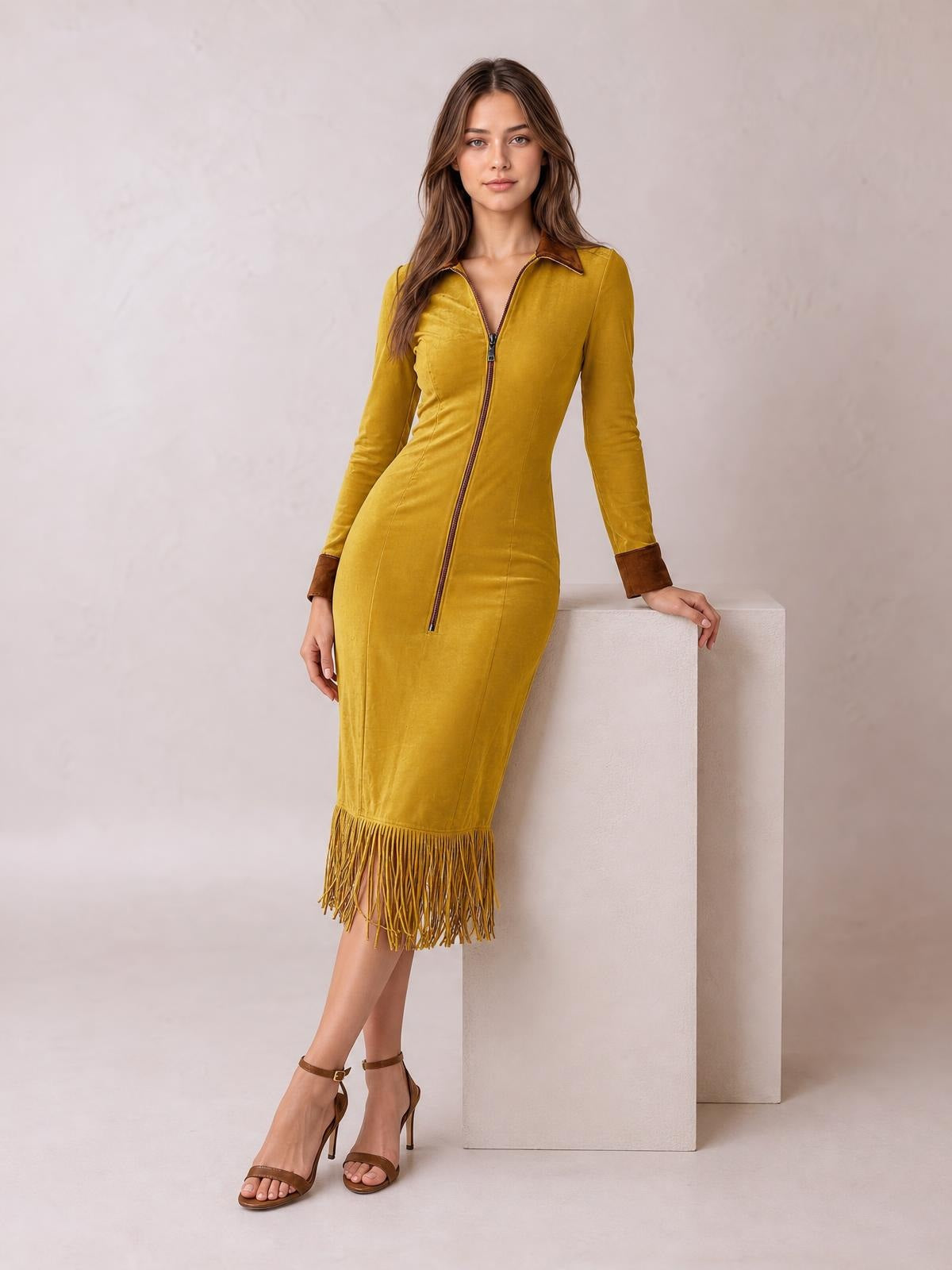 Model wears a mustard yellow fitted midi dress with long sleeves, a zip front, collared neckline, and dramatic fringe hem, posed with one hand on a pedestal.