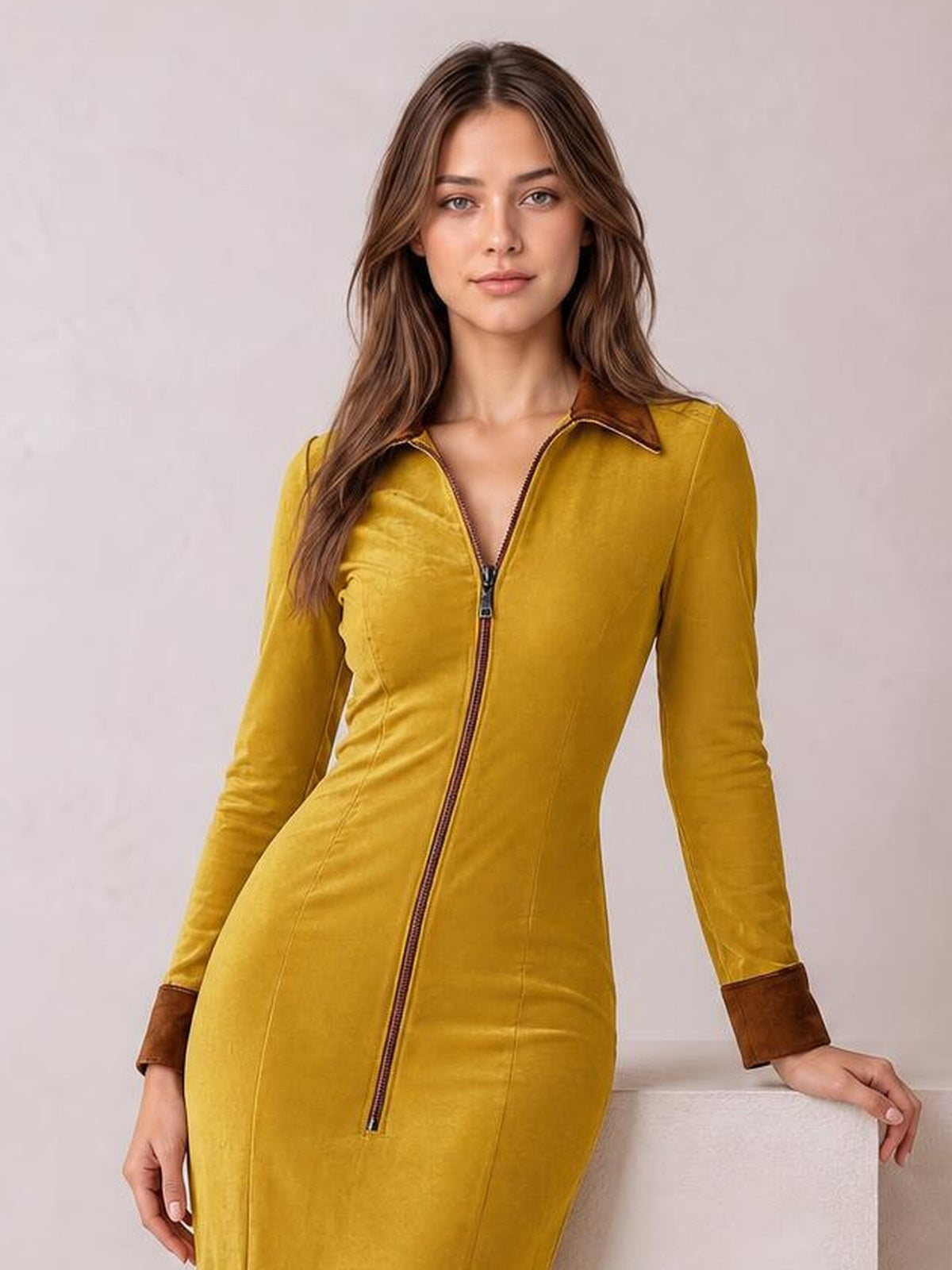 A model poses in a mustard yellow fitted midi dress with a front zipper, pointed collar, long sleeves, brown cuffs, and a streamlined silhouette.
