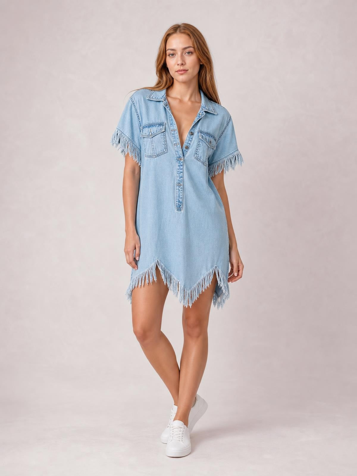 Model stands with crossed legs wearing a light blue denim shirt dress with button front, collared neckline, chest pockets, fringed short sleeves, and uneven fringed hem.