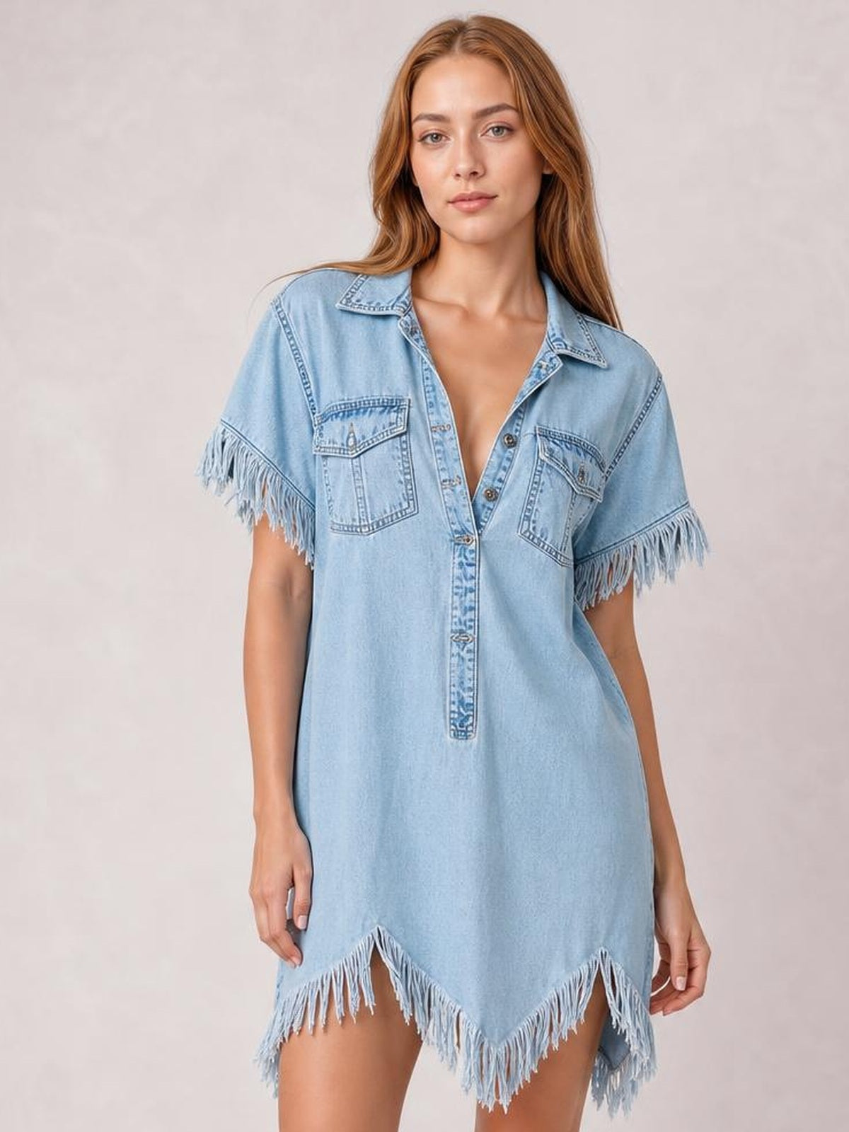 Model stands in a light blue denim shirt dress with a relaxed silhouette, collared button front, chest pockets, fringed short sleeves, and an uneven fringed hem.
