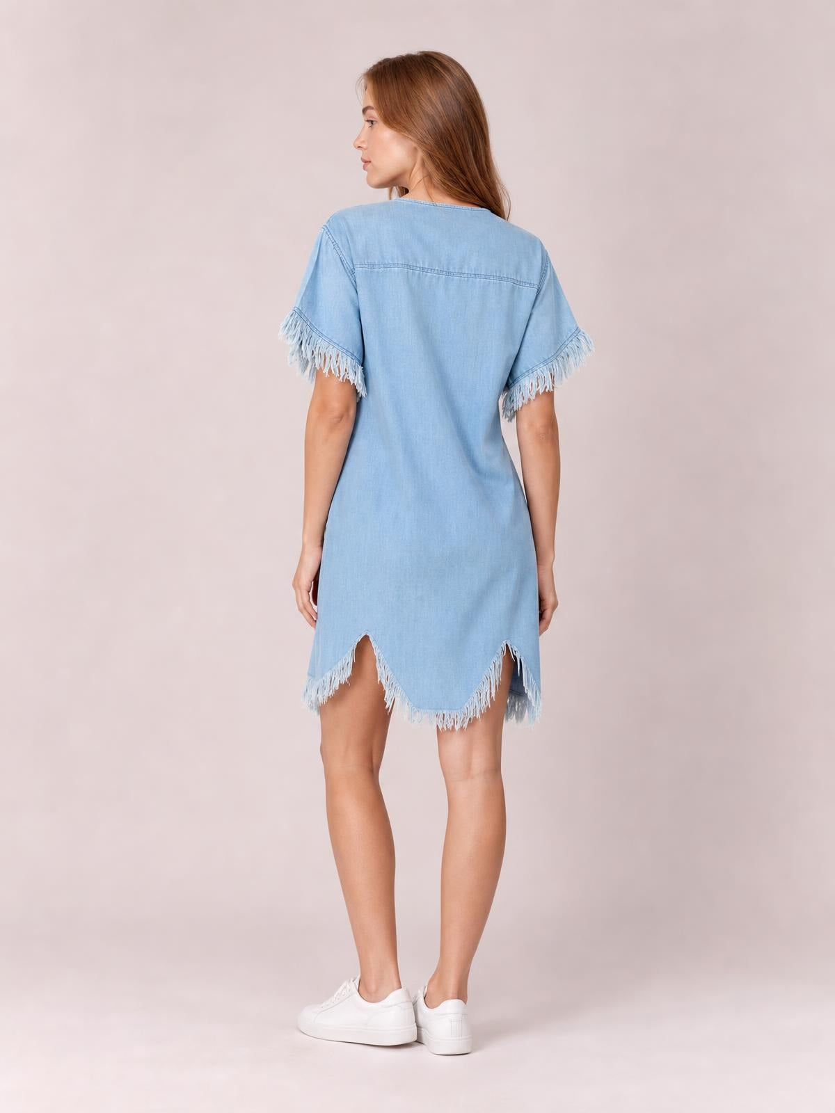 A model stands in a back-facing pose wearing a light blue denim shirt dress with a straight silhouette, fringed short sleeves, and a fringed uneven hem.