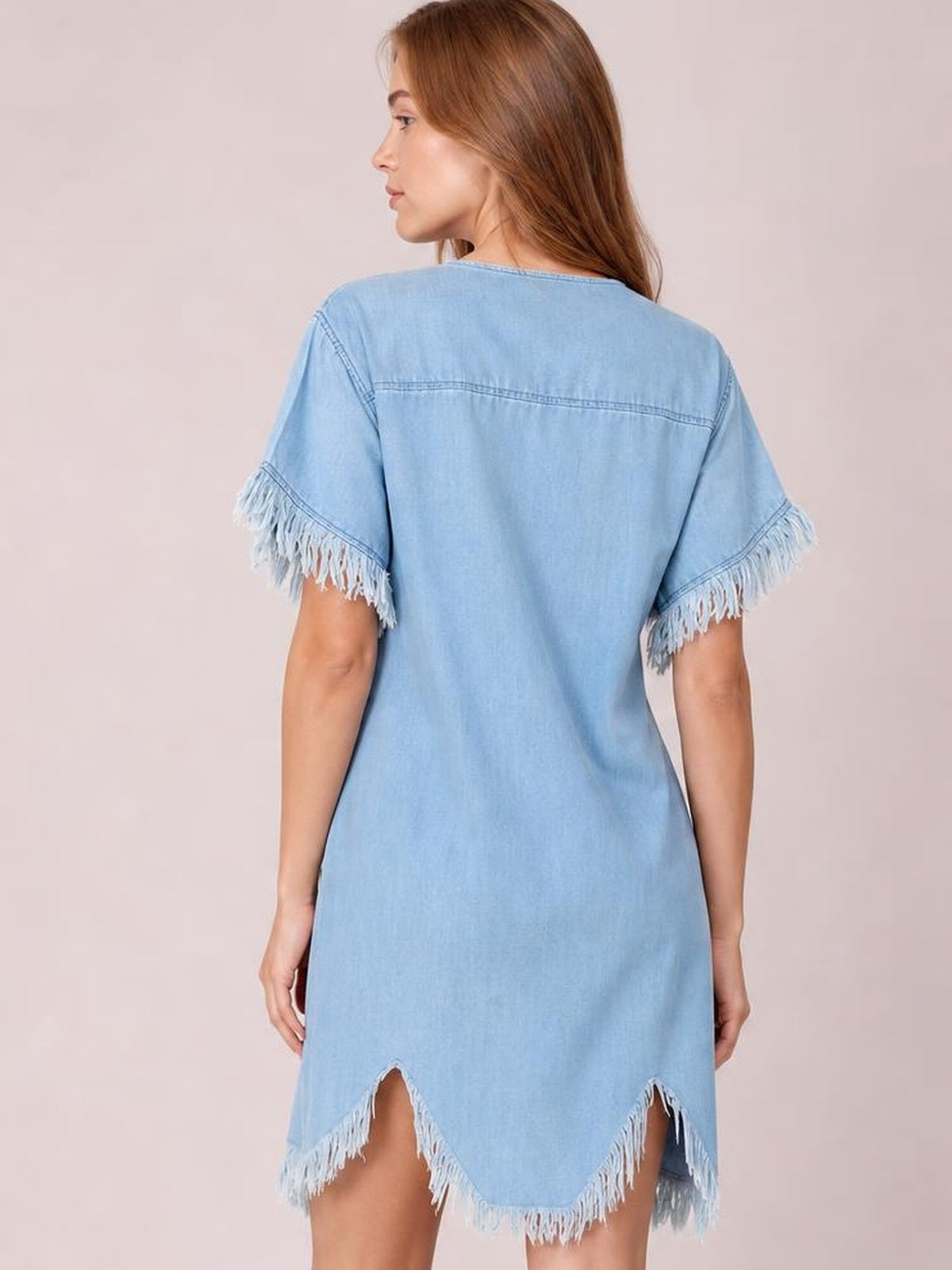 Model shown from behind wearing a light blue denim shirt dress with a relaxed straight silhouette, fringed short sleeves, and an uneven fringed hem.