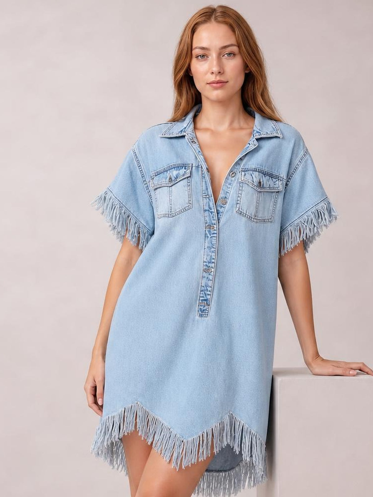 A model stands in a light blue denim shirt dress with a button front, collared neckline, patch pockets, fringed short sleeves and an asymmetrical fringed hem.