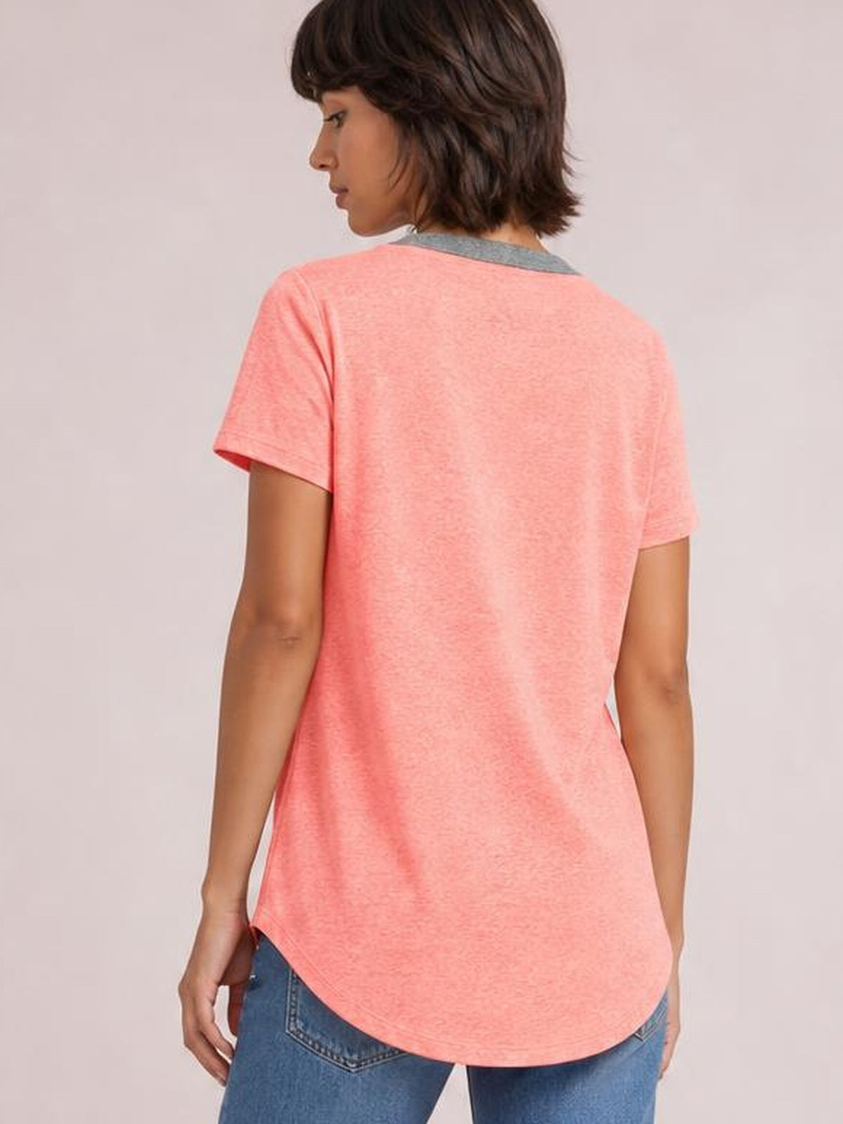 Model turned away in a coral pink short sleeve T-shirt with a relaxed silhouette, rounded shirttail hem, and softly draped back.