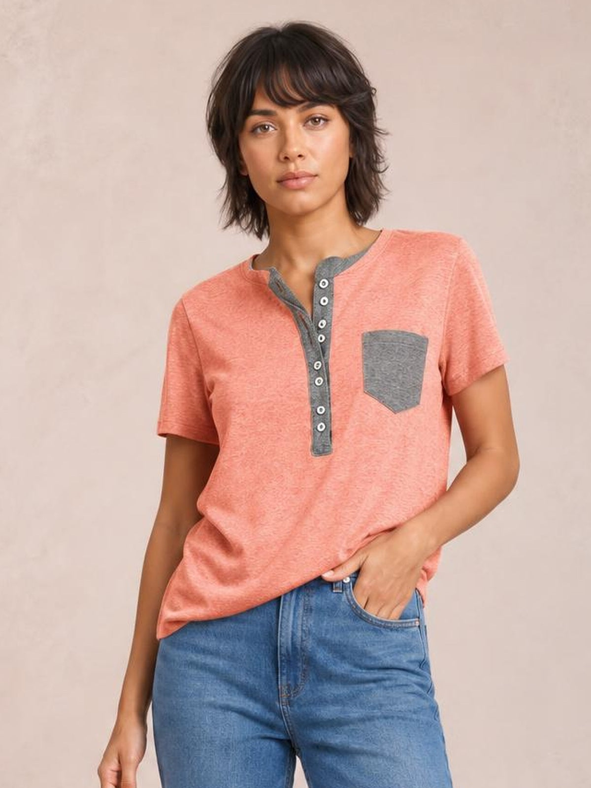 Model wears a coral short sleeve T-shirt with a relaxed silhouette, contrasting gray half-button placket and chest pocket, posed with one hand in her jeans pocket.