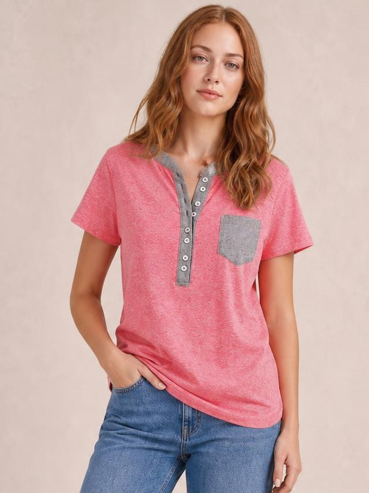 A woman models a relaxed pink short sleeve T-shirt with a gray half-button placket and chest pocket, gently holding the curved hem.