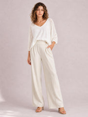 Model poses in cream high-waisted wide-leg pants with front pleats, seam details, and side pockets, styled with a white top.