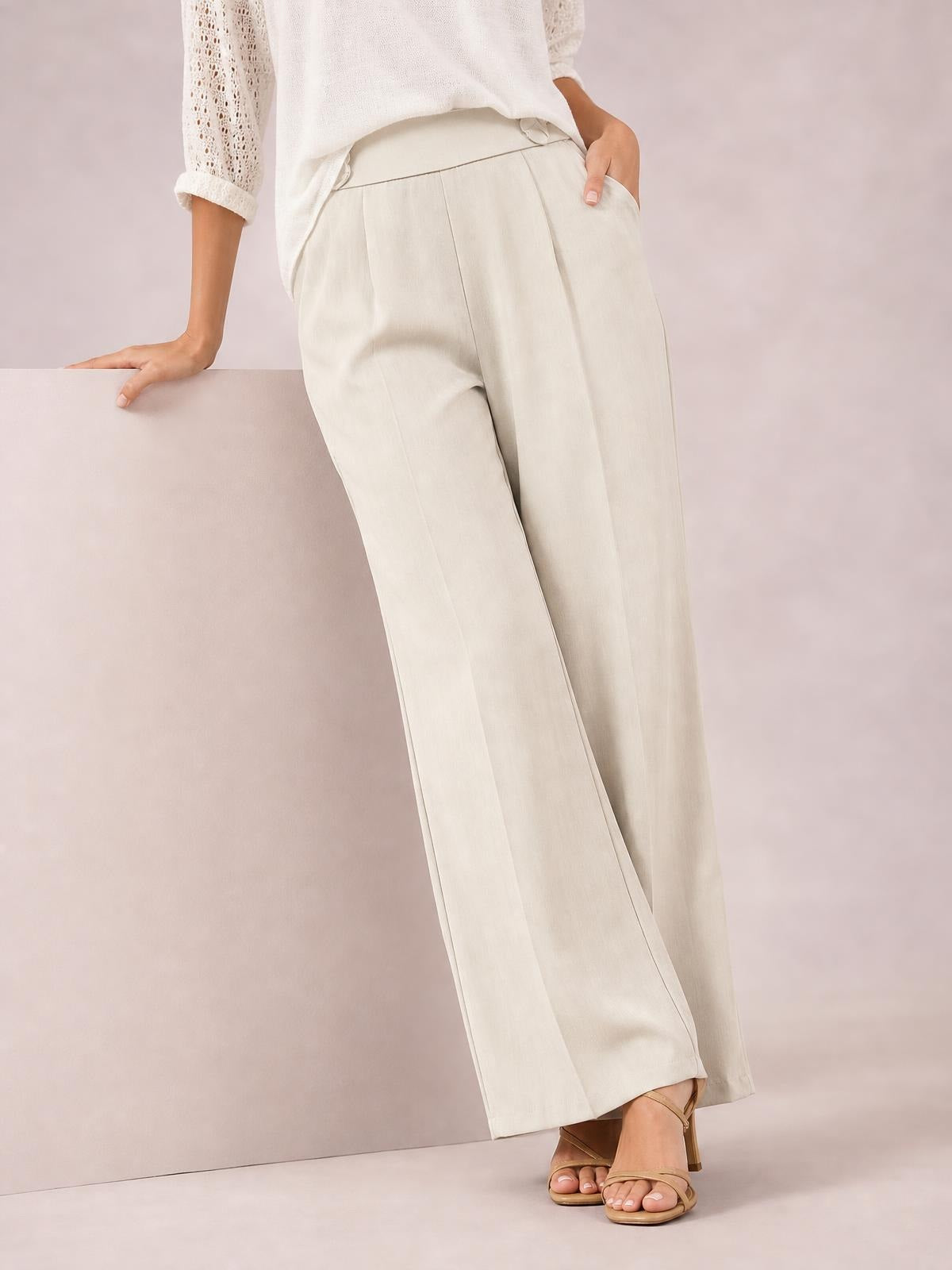 Model wearing cream high-waisted wide-leg pants with front pleats, side pockets, and a relaxed full-length silhouette, standing with one hand in a pocket.