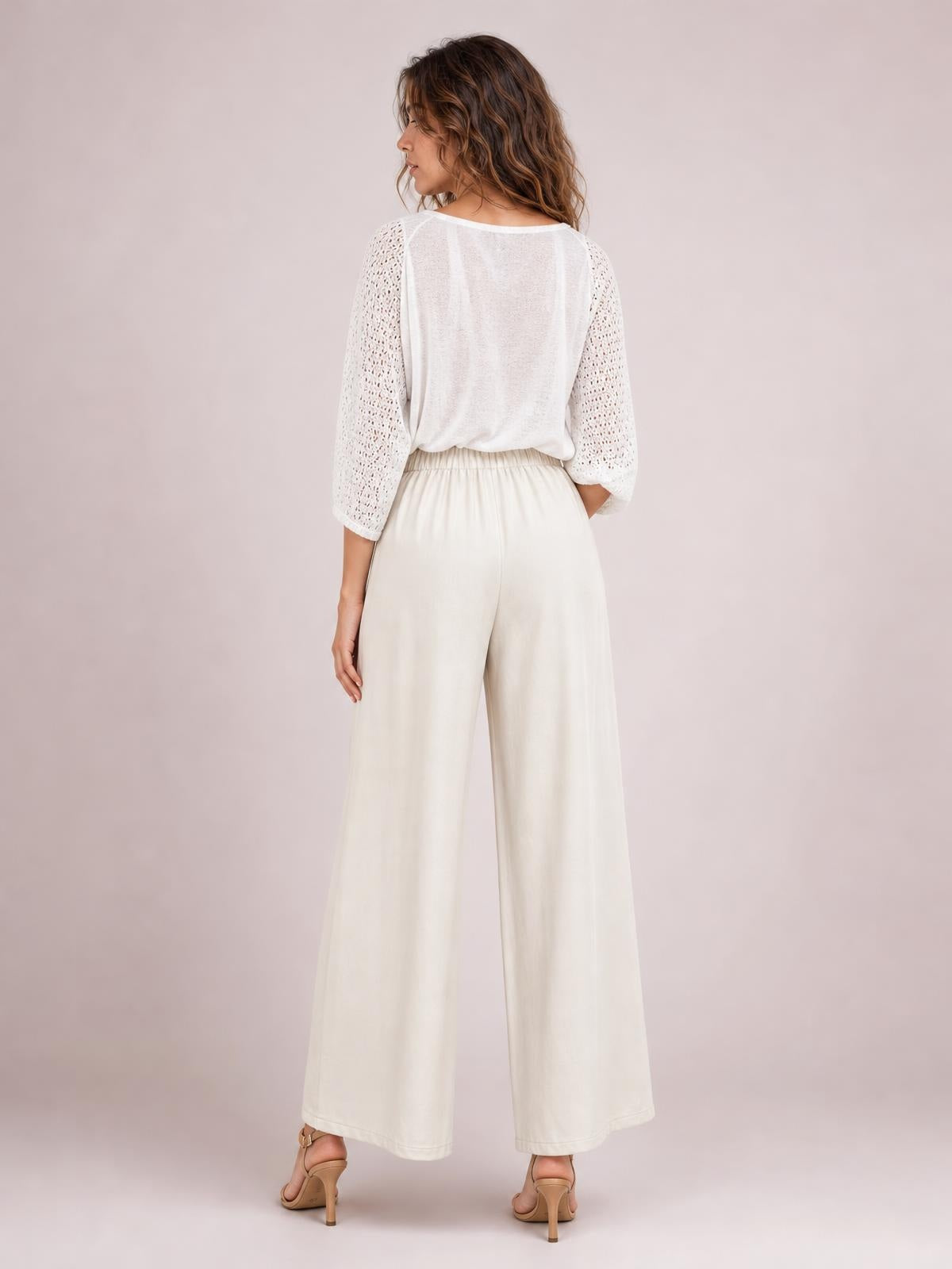 Model shown from the back wearing cream high-waisted wide-leg pants with an elastic waistband, flowing silhouette, and side pockets.