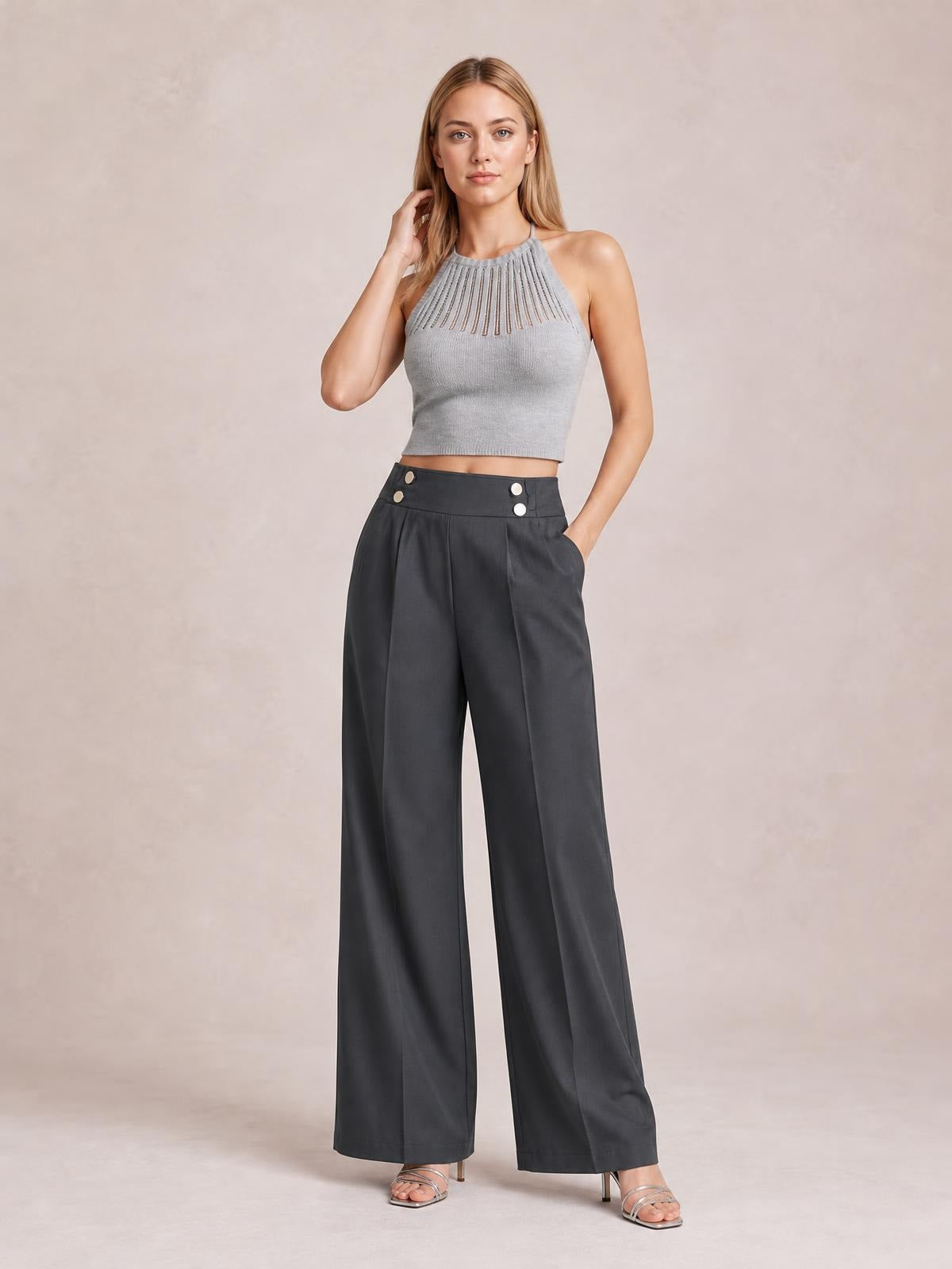 Model wearing charcoal high-waisted wide-leg pants with front pleats, button details, and side pockets, posed with one hand in a pocket.