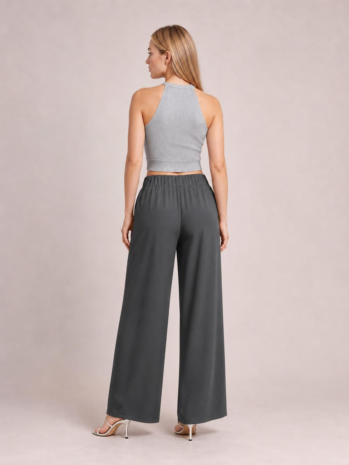 A model stands with her back to the camera in dark charcoal high-waisted wide-leg pants with a gathered waistband and full-length silhouette.